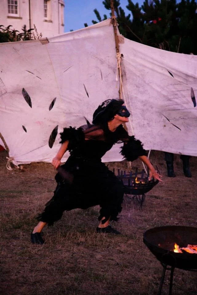 Person dressed in a black costume with feathered details, wearing a hat, dancing or performing in front of a makeshift canvas backdrop during an outdoor evening event with fire pits.