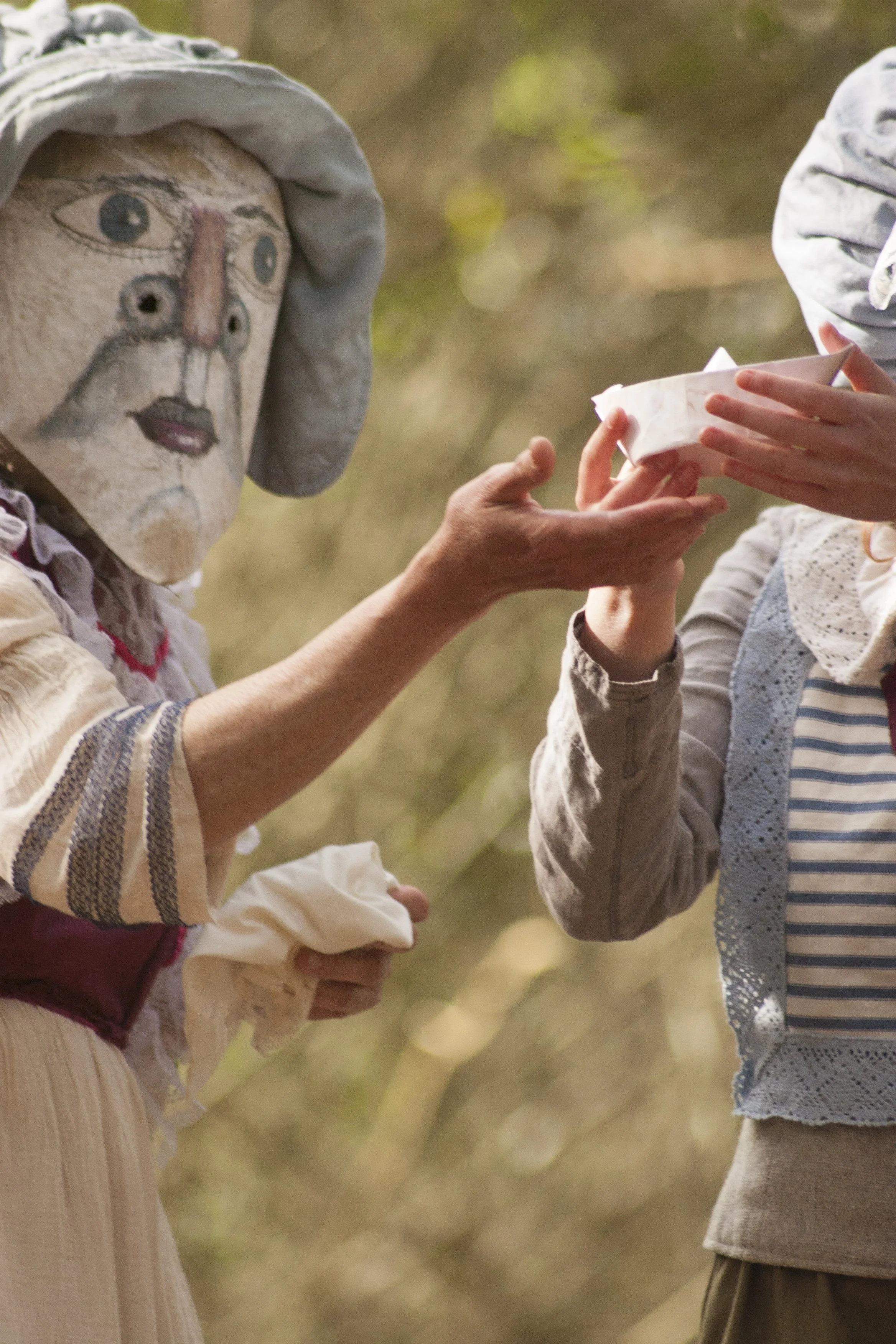 Two individuals exchanging a paper container outdoors, one with a painted face mask resembling a human face with multiple eyes, nose, and mouth, wearing a striped shirt and cream-colored skirt, the other with a strappy dress and lace vest, holding th