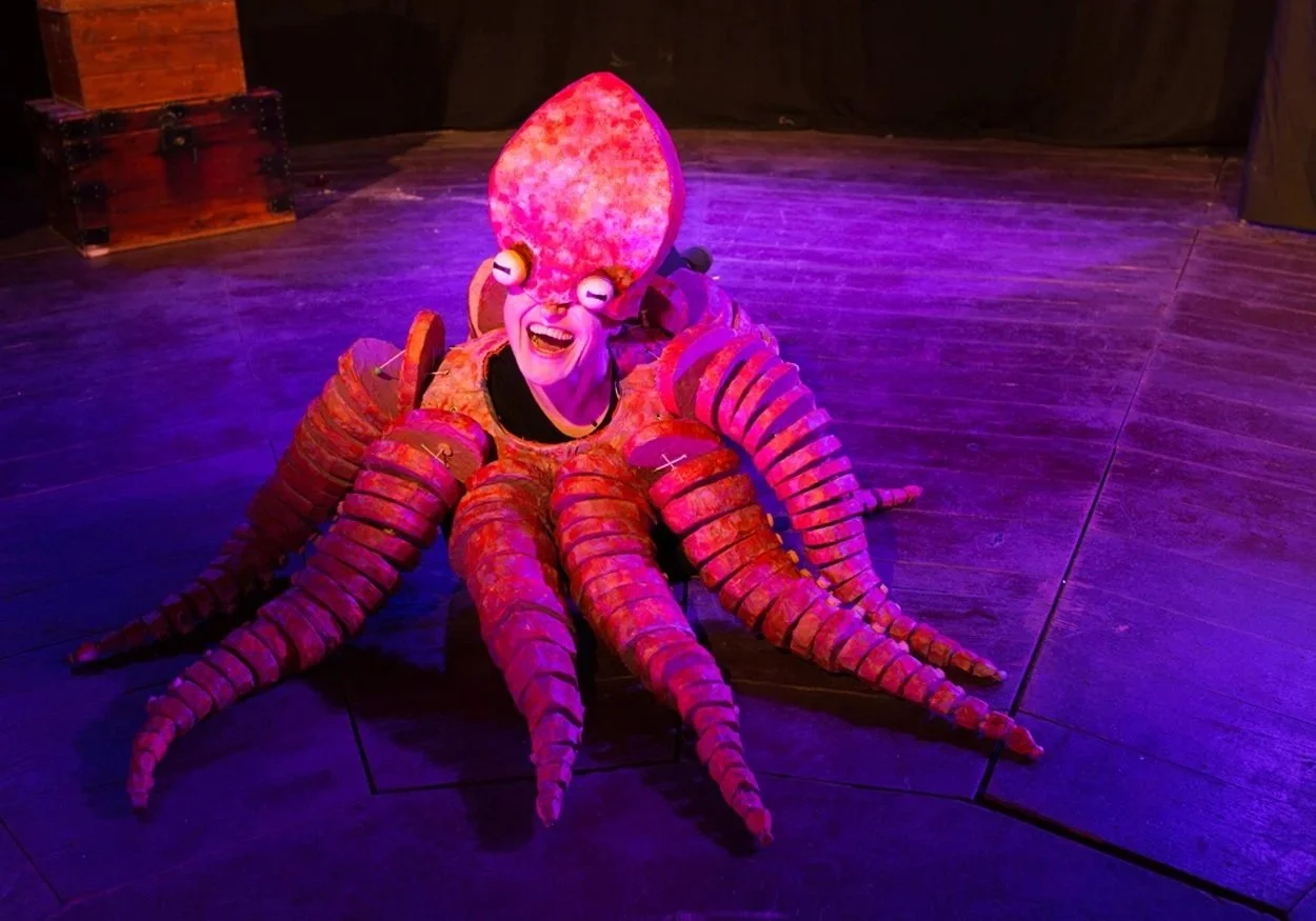 A person dressed as a bright pink and orange octopus with a large head and multiple tentacles, smiling and posing on a dark wooden floor with purple lighting.