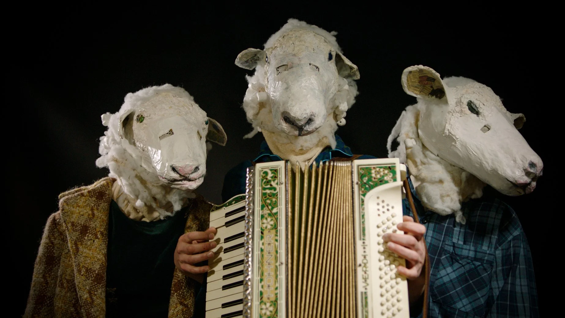 Three individuals wearing sheep masks and vintage clothing, playing musical instruments on stage, with a dark background.