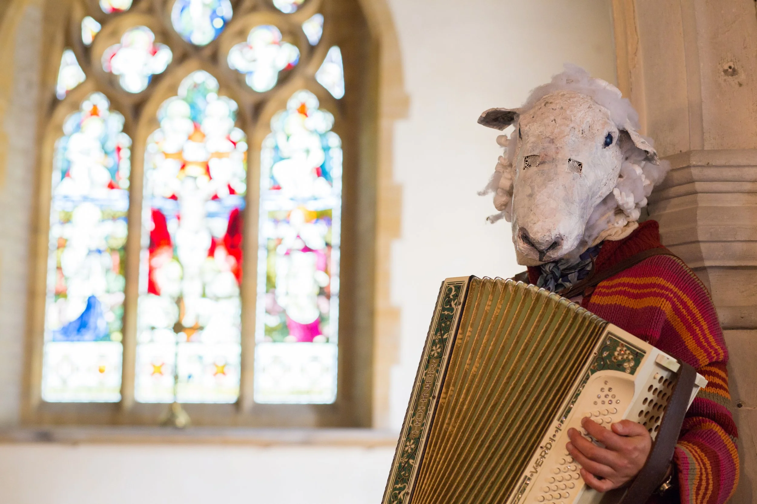 A person wearing a sheep mask playing an accordion inside a church or cathedral, with stained glass windows in the background.