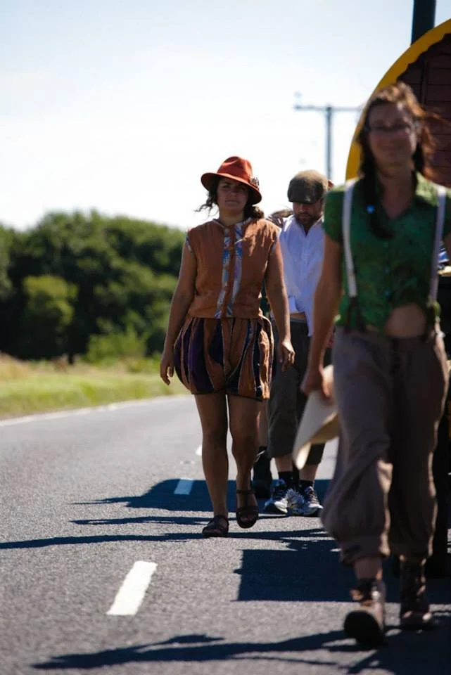 Three people walking on a road with green trees in the background, including a woman in a brown hat and patterned outfit.