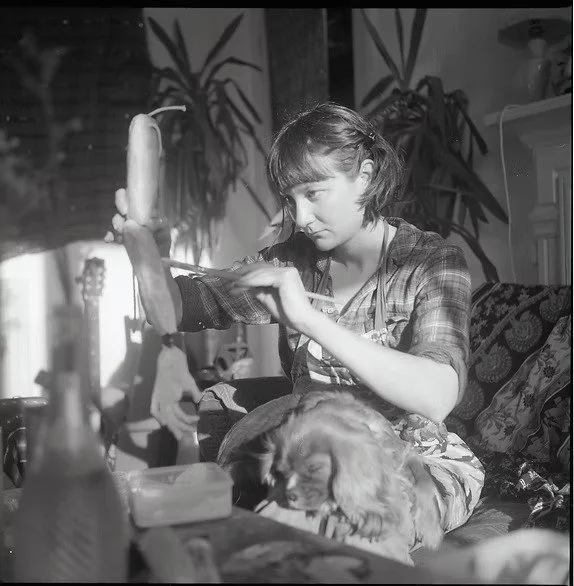 A woman sits on a couch making a puppet, in a room with plants and guitars in the background.