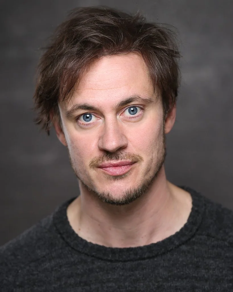 Close-up portrait of a young man with blue eyes, brown hair, light facial hair, and wearing a dark sweater, against a dark gray background.