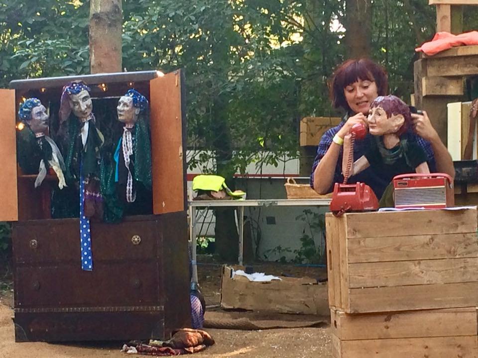 A woman performs with puppets outdoors, using a telephone puppet to speak to a puppet on the table. A puppet theater with three marionettes is visible on the left.