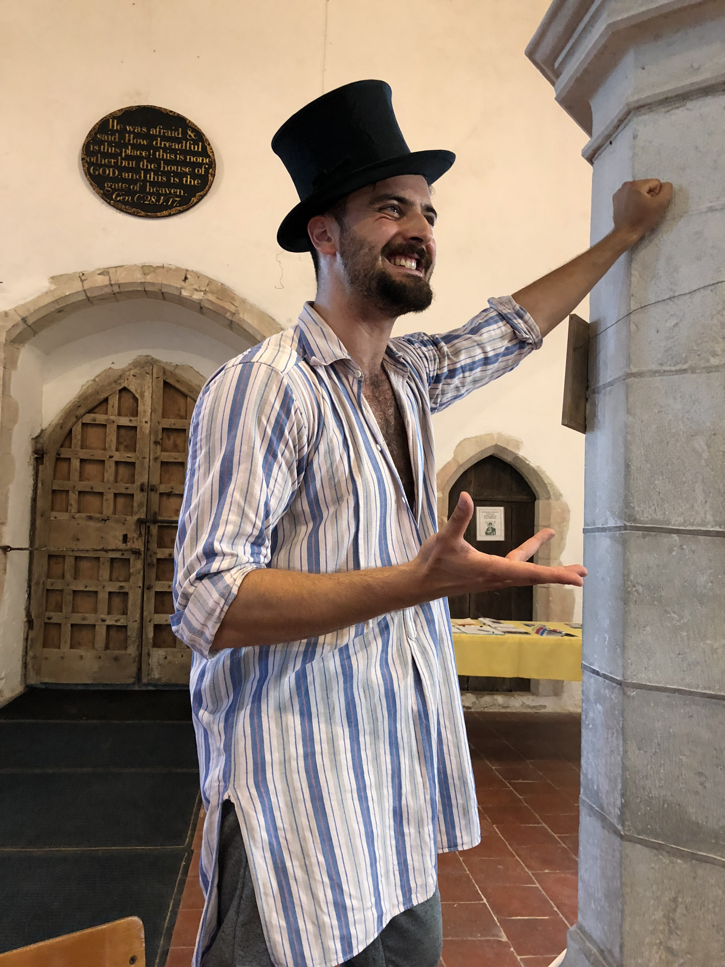 A man with a beard wearing a top hat and striped shirt smiling and gesturing with his hands indoors, with a stone pillar on his right and a wooden door in the background.