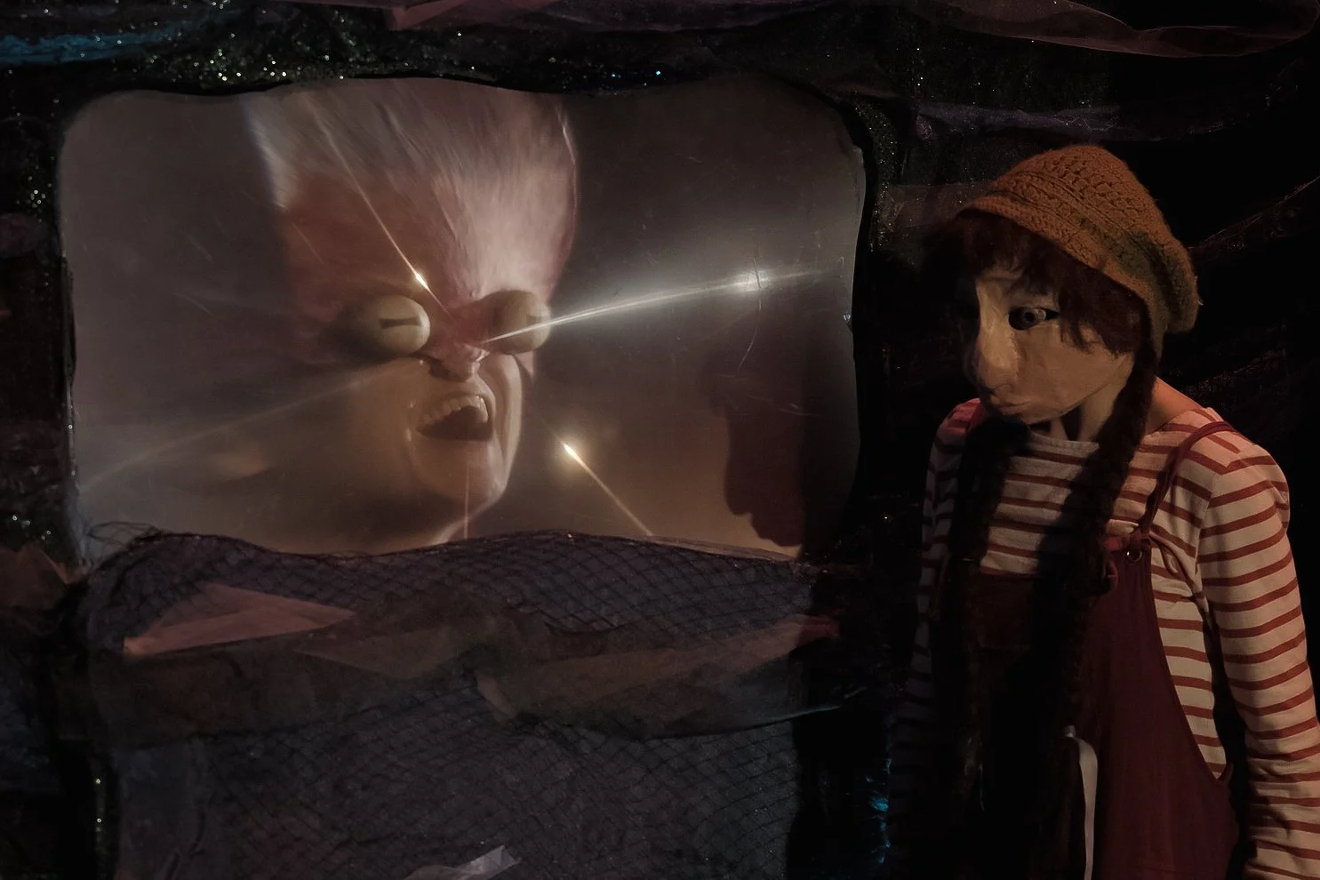 A girl with braided hair and a striped shirt wearing a brown knit cap, looking at a ventriloquist dummy inside a glass enclosure, which has exaggerated facial features and appears to be angry or distressed.