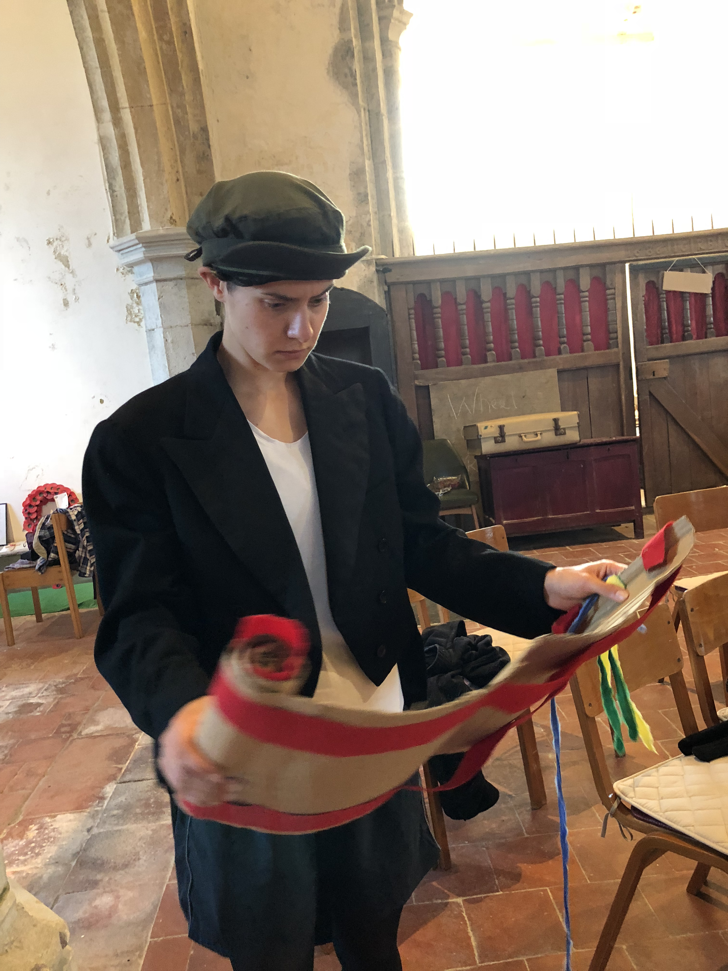 A young person wearing a gray cap and black jacket, looking down at and reading a booklet, in a room with stone walls and wooden chairs, with a bright window in the background.