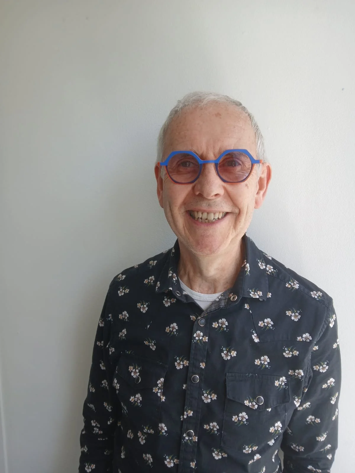 A smiling elderly man with gray hair wearing fashionable blue and red sunglasses and a black shirt with white daisy patterns, standing against a plain white wall.