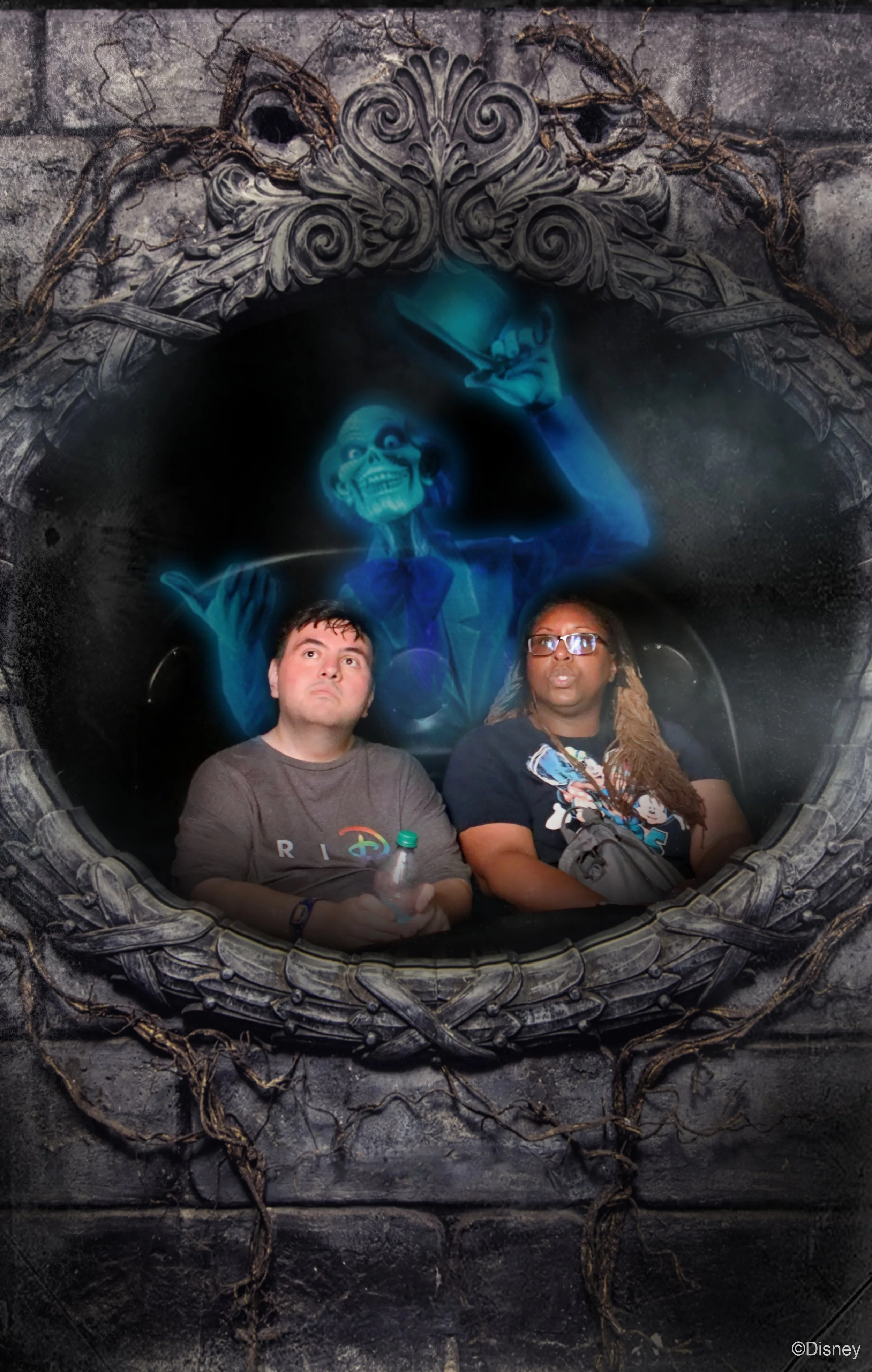 Two people sitting in front of a Disney-themed haunted house display with a glowing blue ghostly figure of a man in a suit, surrounded by dark, weathered architecture and vines.