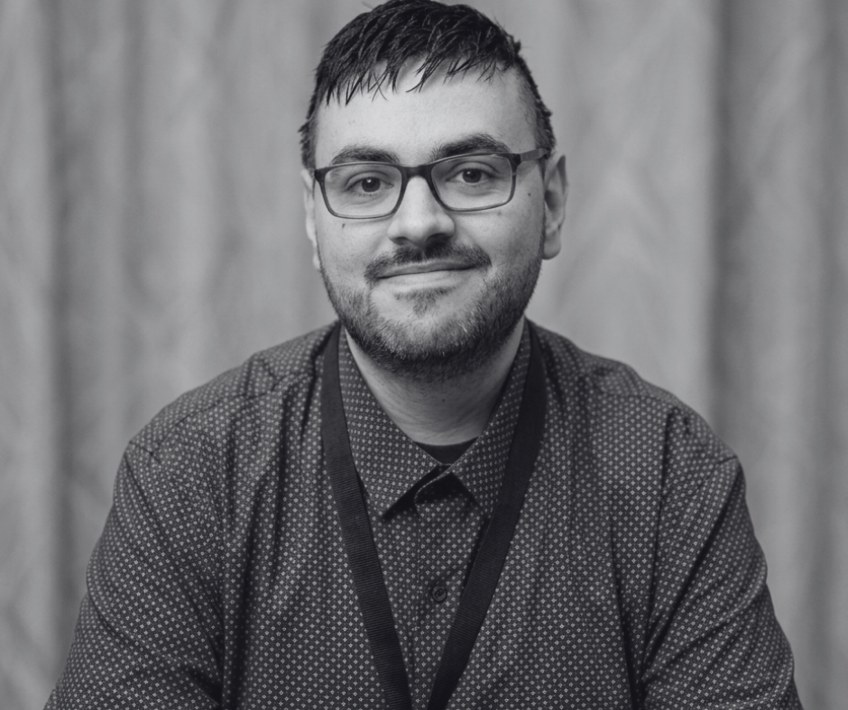 A man wearing glasses and a button-up shirt, smiling at the camera with a plain background.