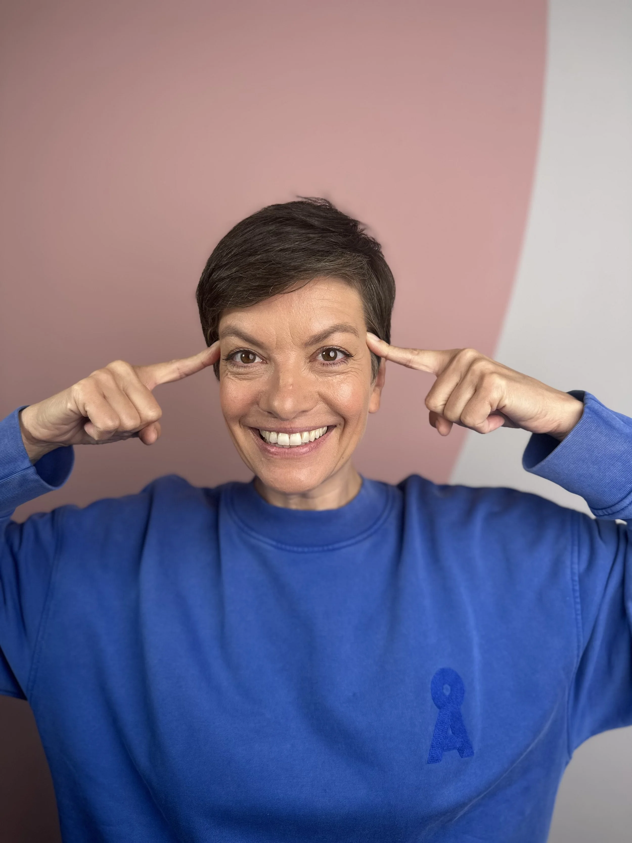 Eine lachende Frau mit kurzen dunklen Haaren trägt ein blaunes Sweatshirt mit einem blauen Aufnäher und berührt ihre Schläfen mit den Fingern vor einem zweifarbigen Hintergrund in Rosa und Weiß.