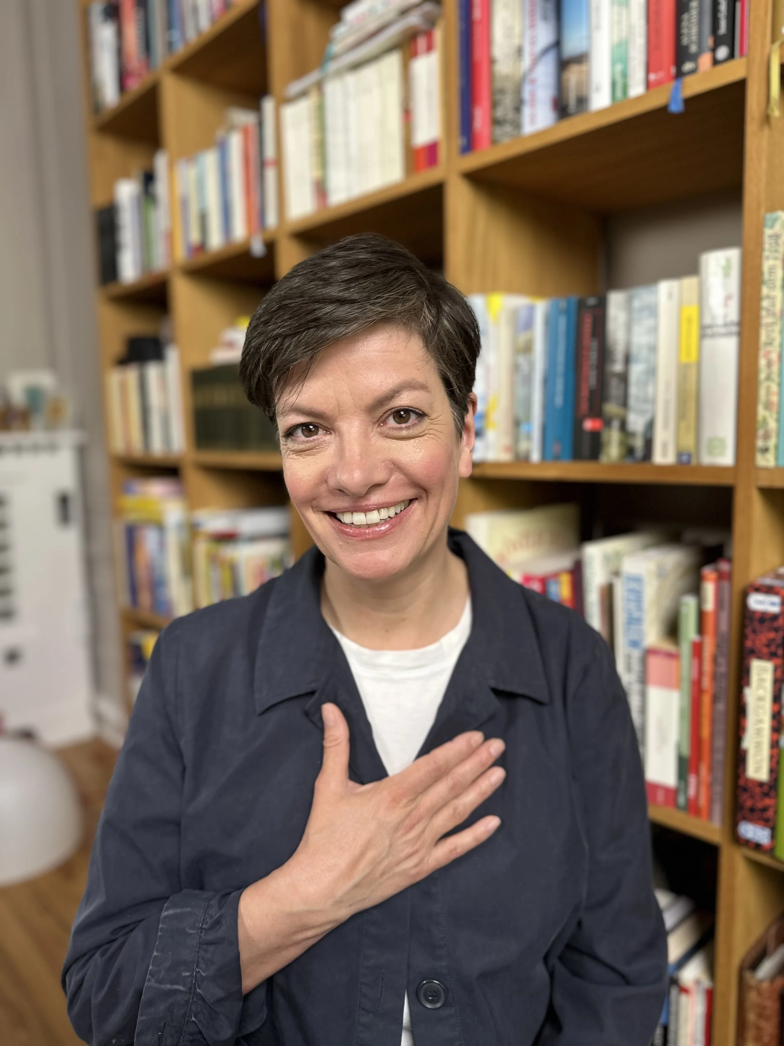 Eine lachende Frau mit kurzen braunen Haaren, trägt eine schwarze Jacke und einen weißen T-Shirt, steht in einem Bücherregal, hält die rechte Hand auf die Brust, die Bücher im Hintergrund.