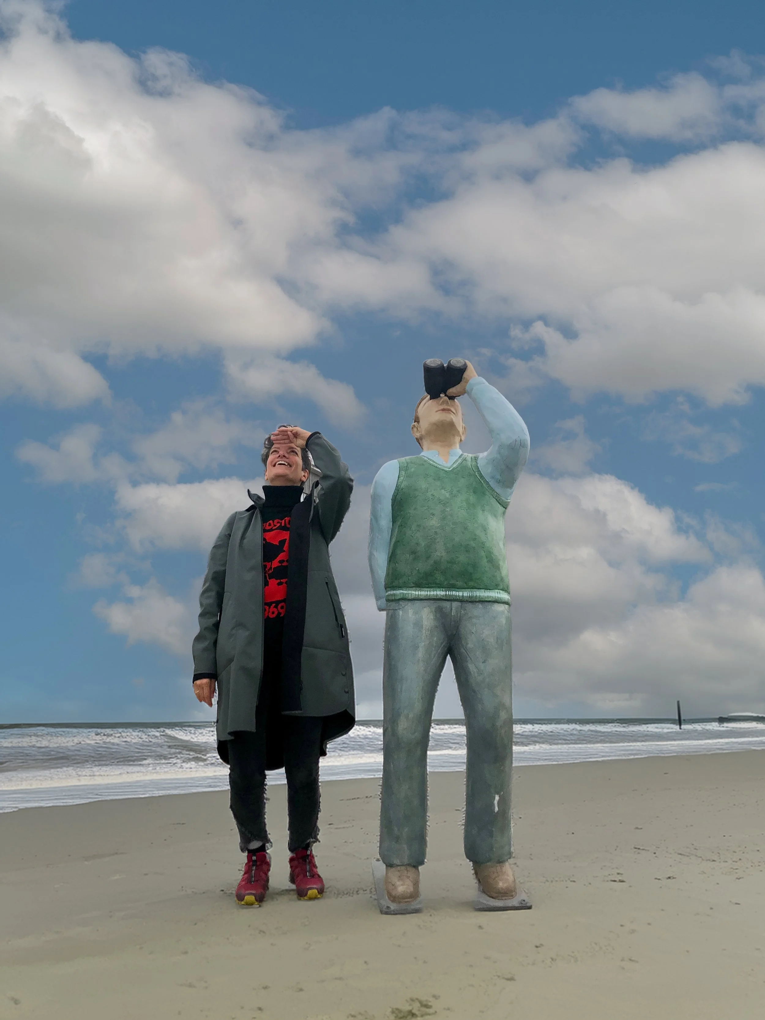 Eine Frau steht neben einer Skulptur eines Mannes, der mit einem Fernglas in den Himmel schaut, am Strand bei bewölktem Himmel.