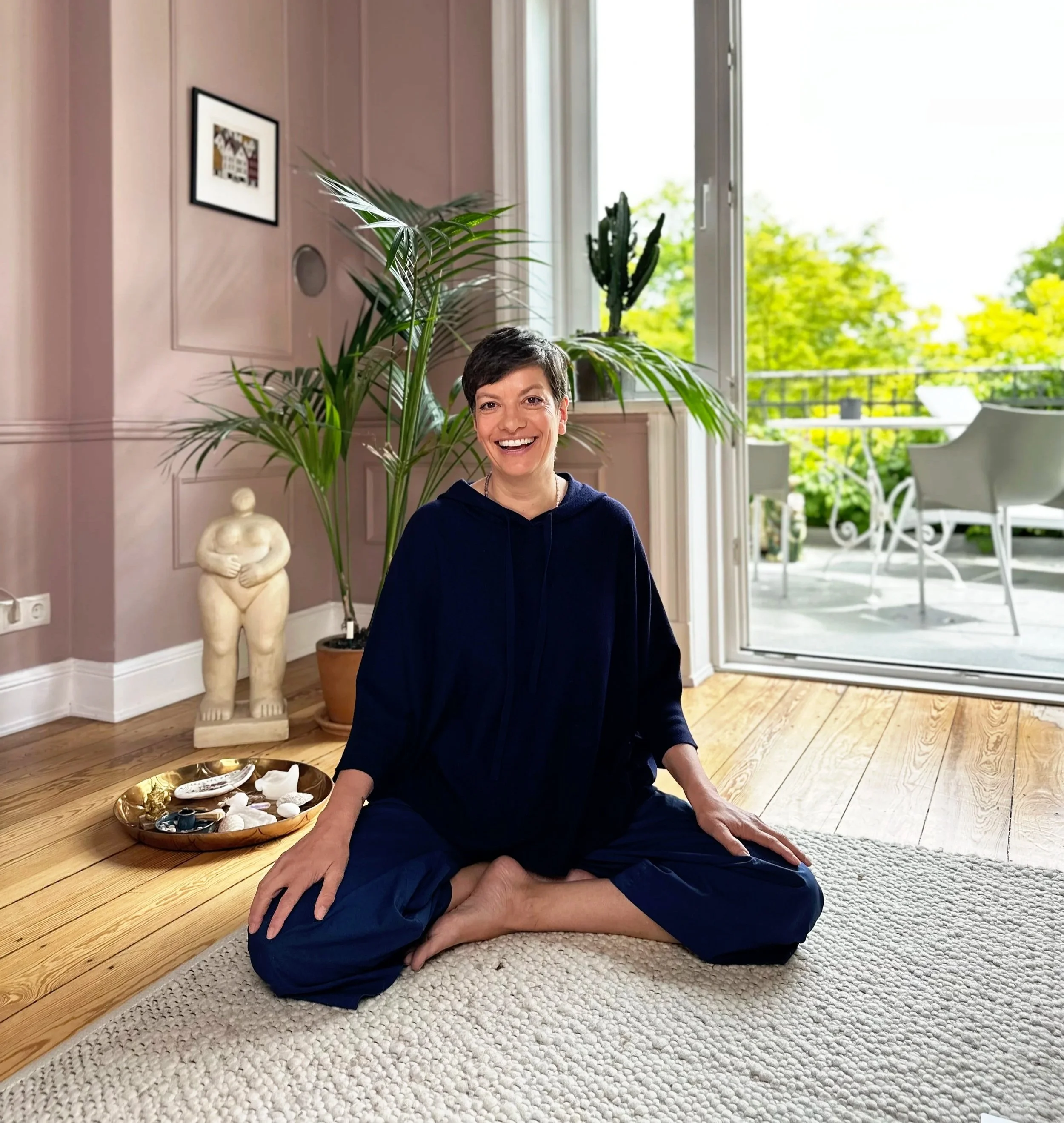 Eine fröhliche Frau sitzt im Schneidersitz auf einem Teppich in einem hell erleuchteten Raum mit offenen Türen, die auf eine Terrasse mit Gartenblick führen.