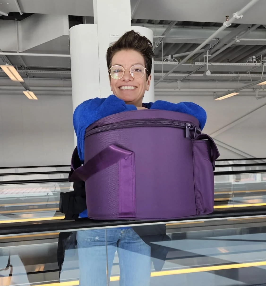 Lächelnde Frau mit Brille, die einen großen, lila Rucksack auf einer Gepäckförderband in einem Flughafen trägt.