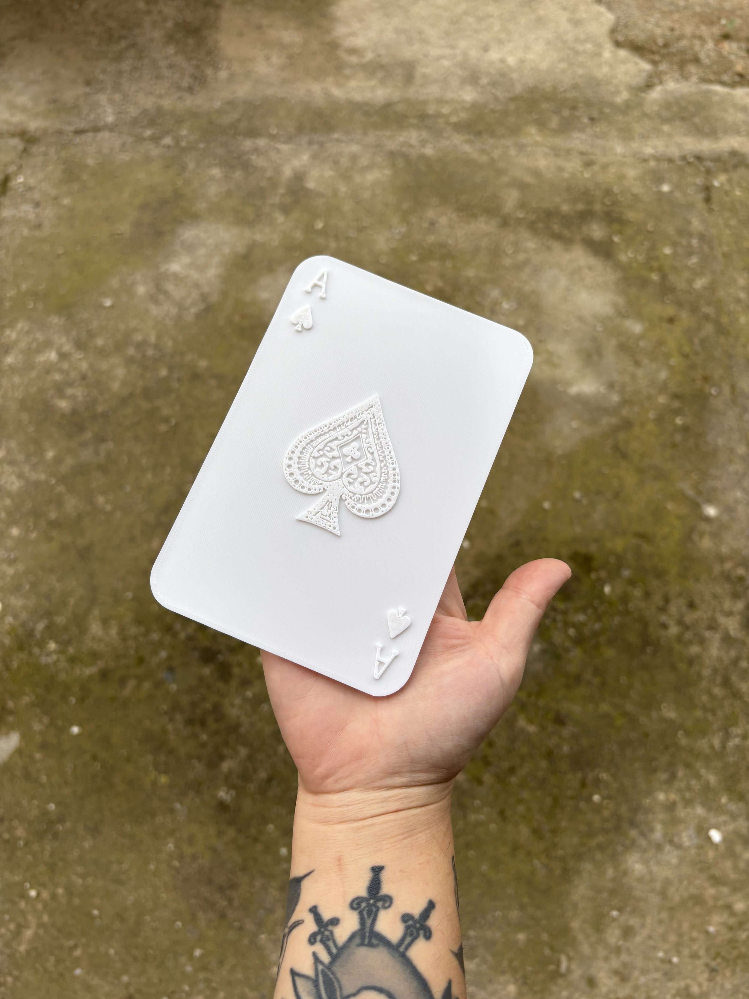 Hand holding a large white deck of playing cards with intricate spade symbol on the front, outdoors on a dirt surface.