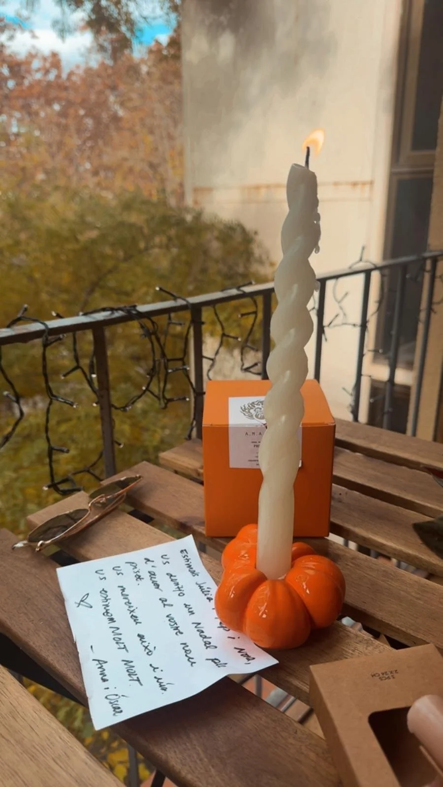 A white twisted candle in a small orange pumpkin-shaped holder on a wooden table with a handwritten note, sunglasses, and an orange box in the background, with a balcony railing and autumn trees outside.