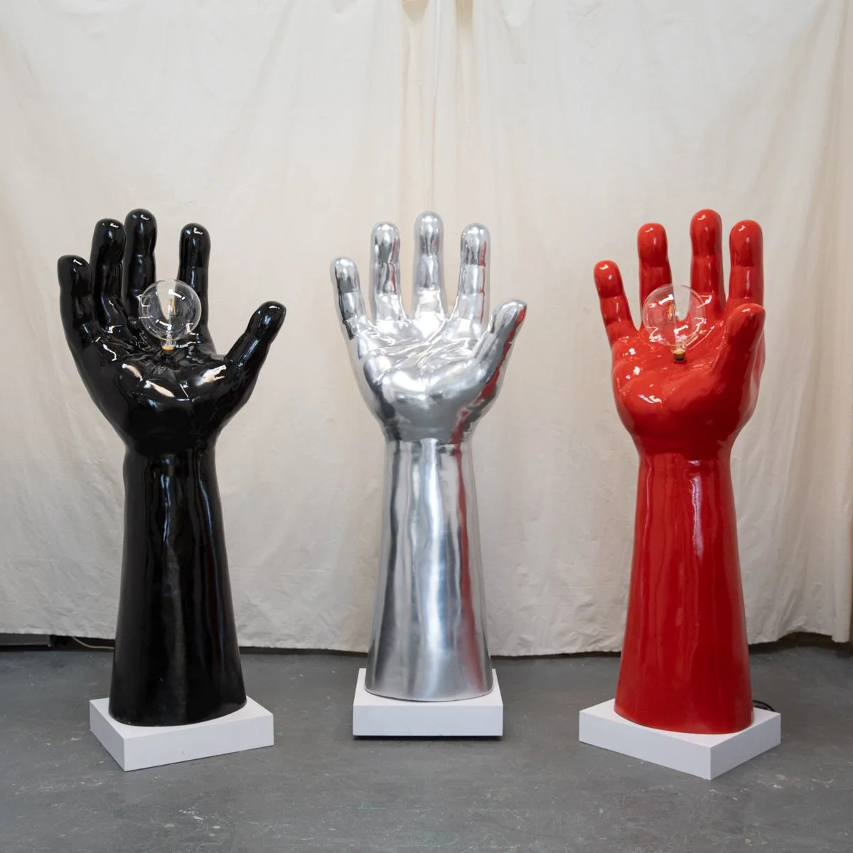 Three large decorative hand sculptures in black, silver, and red, each with a light bulb in the palm, displayed on white bases against a cream-colored background.