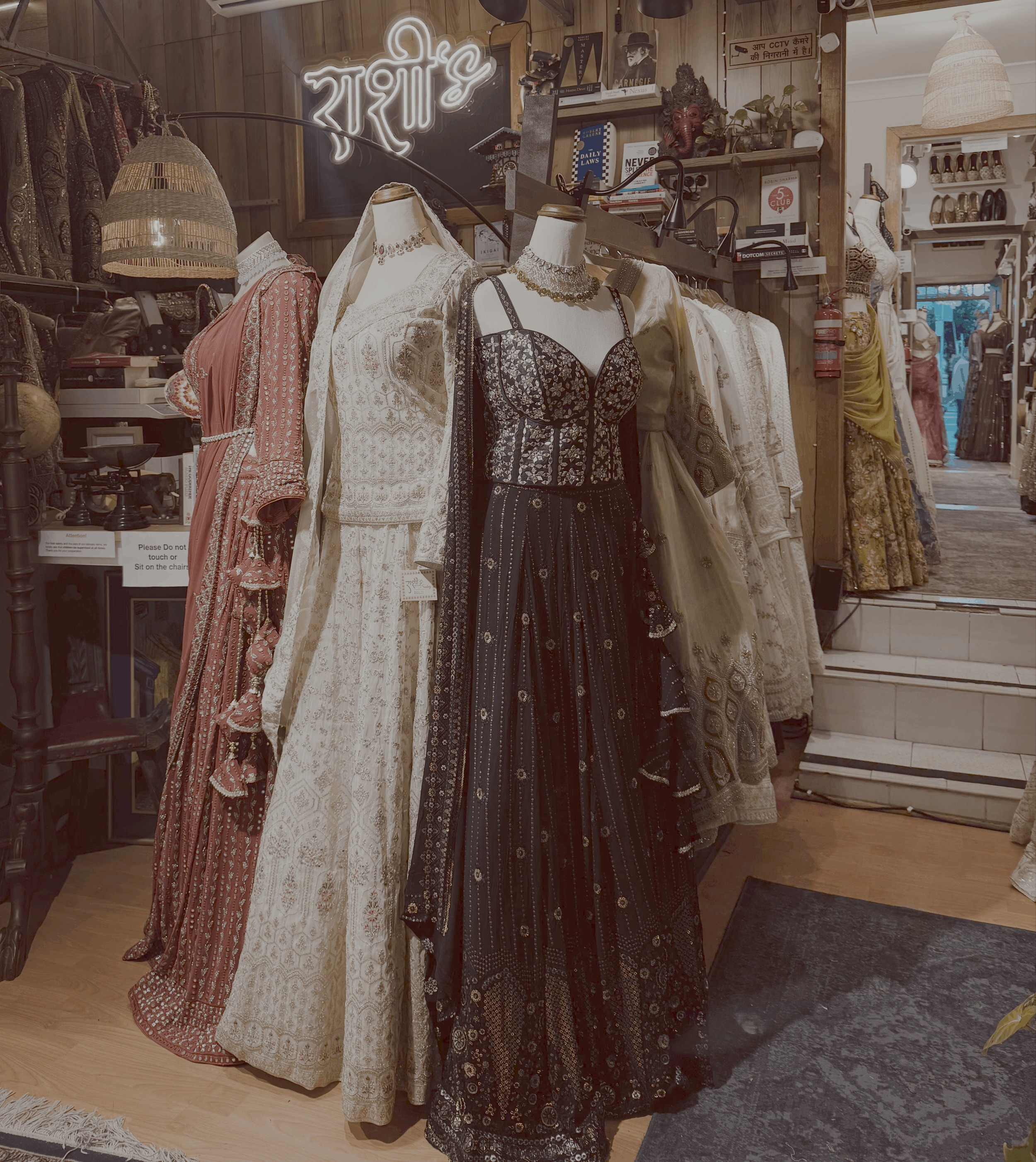 Display of traditional ethnic dresses and jewelry on mannequins inside a boutique or clothing store, with wooden decor and books on shelves, and a neon sign in Hindi language.