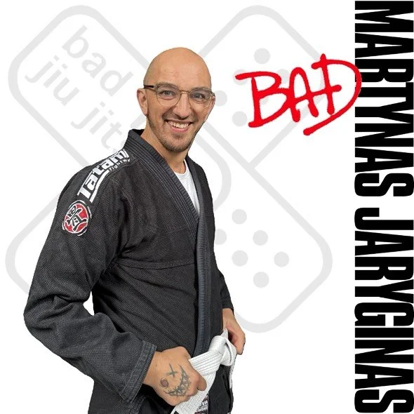 Man in Brazilian Jiu Jitsu gi standing smiling with a white belt, background with the words 'BAD' in red and 'MARTYNAS JARYCNAS' in black.