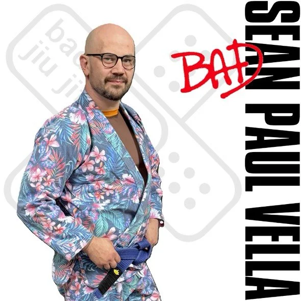 A man wearing glasses and a floral-patterned judo gi with a blue belt, standing with one hand in his pocket, against a backdrop with black text and a red graffiti-style word 'BAD'.