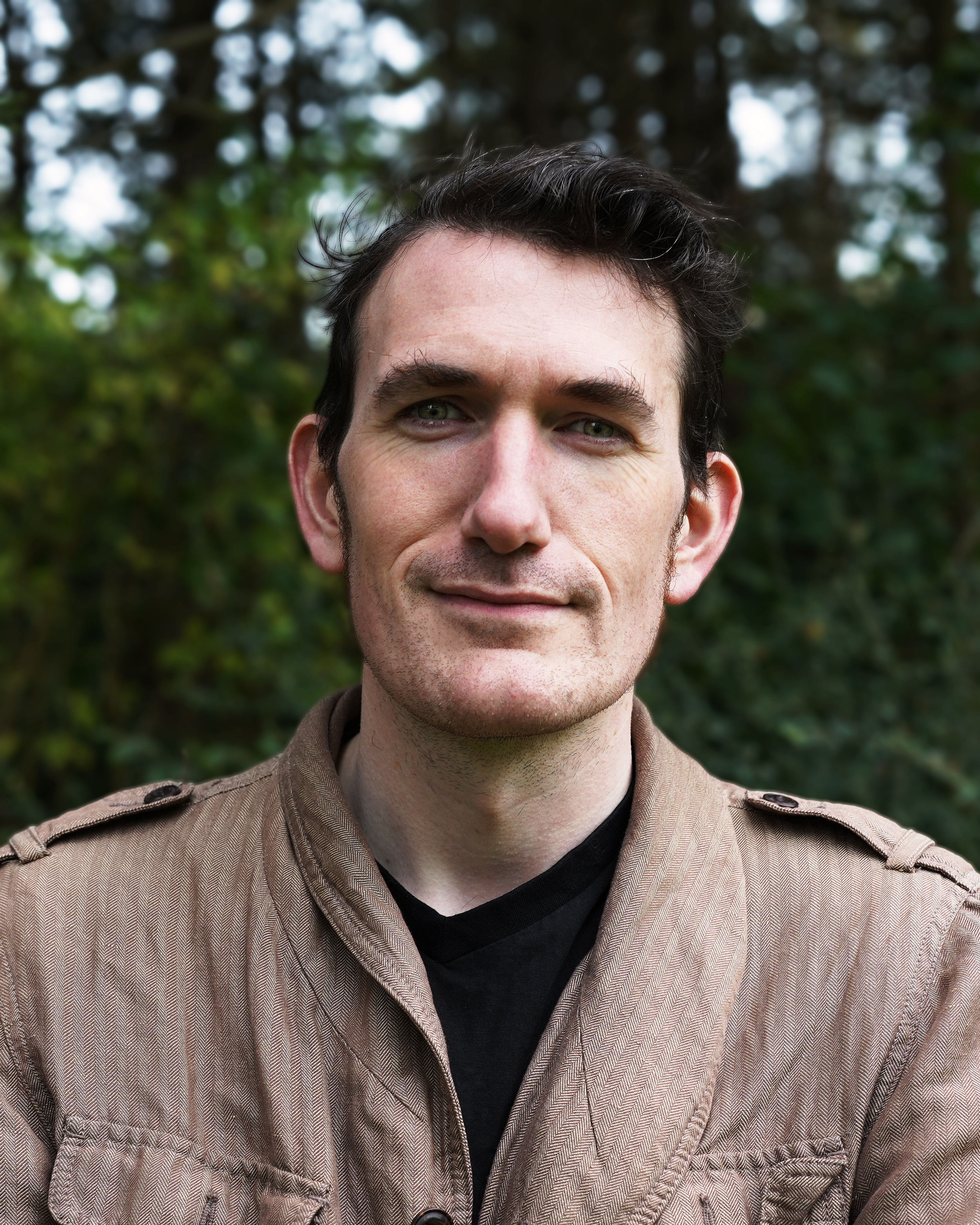 A man with short dark hair and light skin standing outdoors with trees in the background.