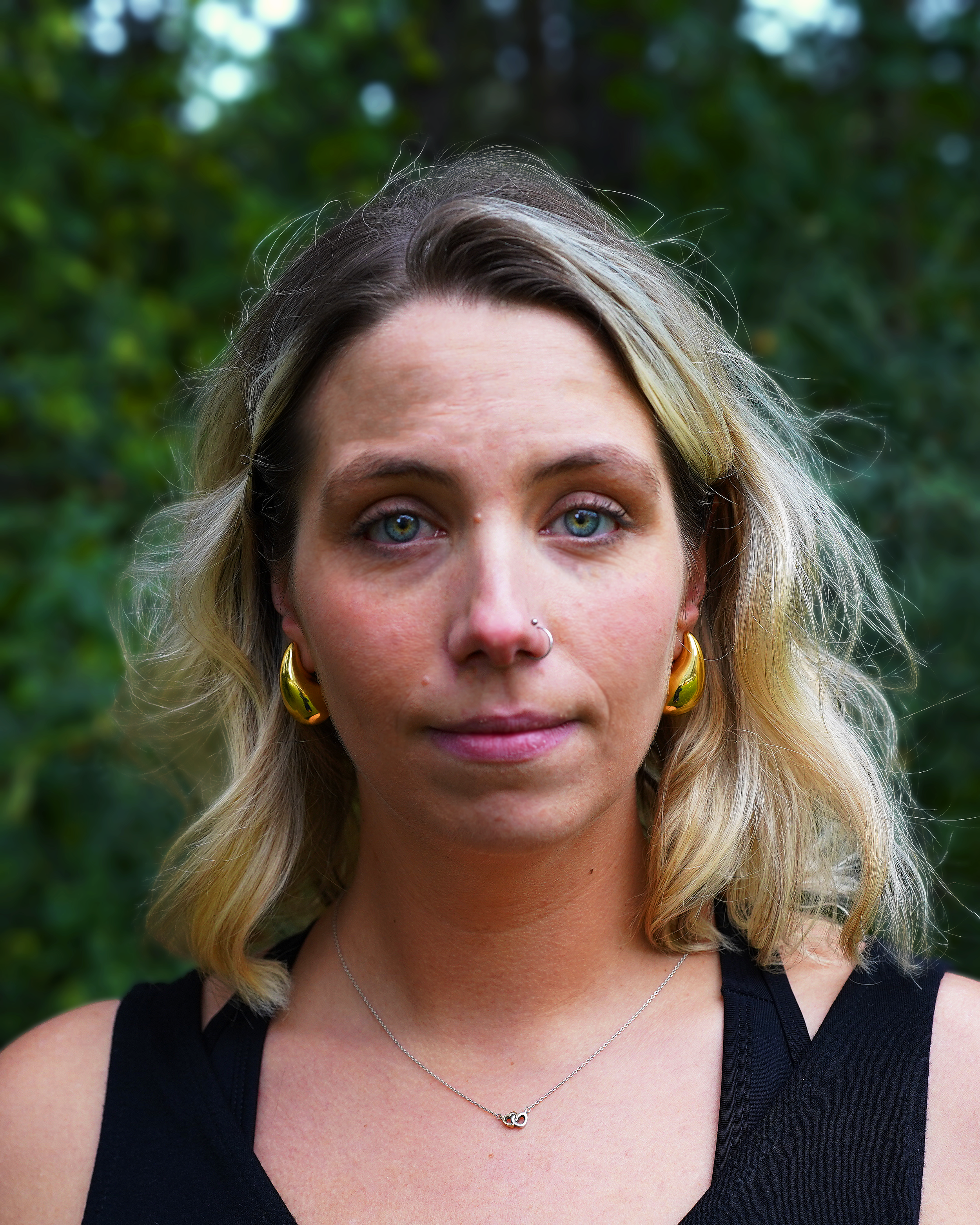 Close-up of a woman with shoulder-length blonde hair, blue eyes, gold hoop earrings, a nose ring, and a silver necklace, standing outdoors with a blurred green foliage background.