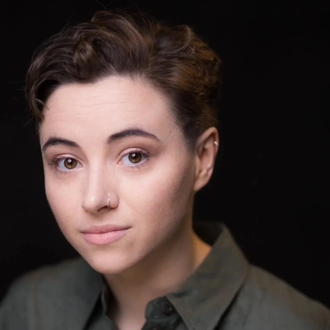 Close-up portrait of a young woman with short, dark hair, brown eyes, wearing a dark green shirt, and a nose ring, against a black background.