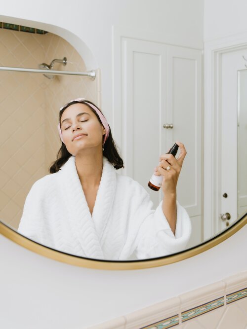 A young woman in a white bathrobe and pink headband sprays a facial mist in front of a mirror in a bathroom.
