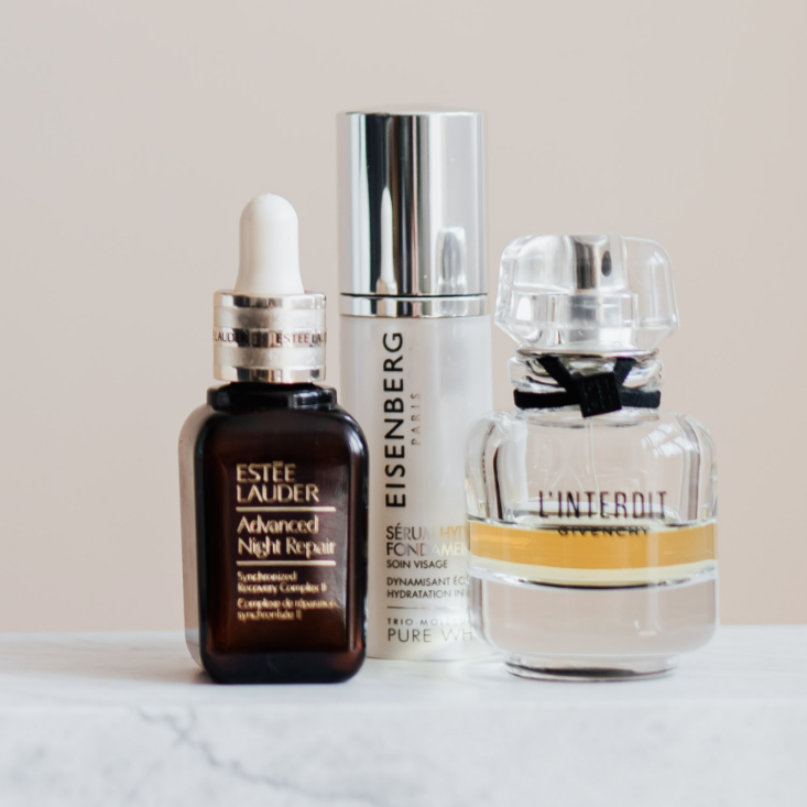 Three skincare products on a white surface against a plain beige background: a dark brown Estee Lauder Advanced Night Repair serum with a dropper, a white and silver Eisenberg serum in a pump bottle, and a clear Interdit perfume bottle with a gold band and clear cap.