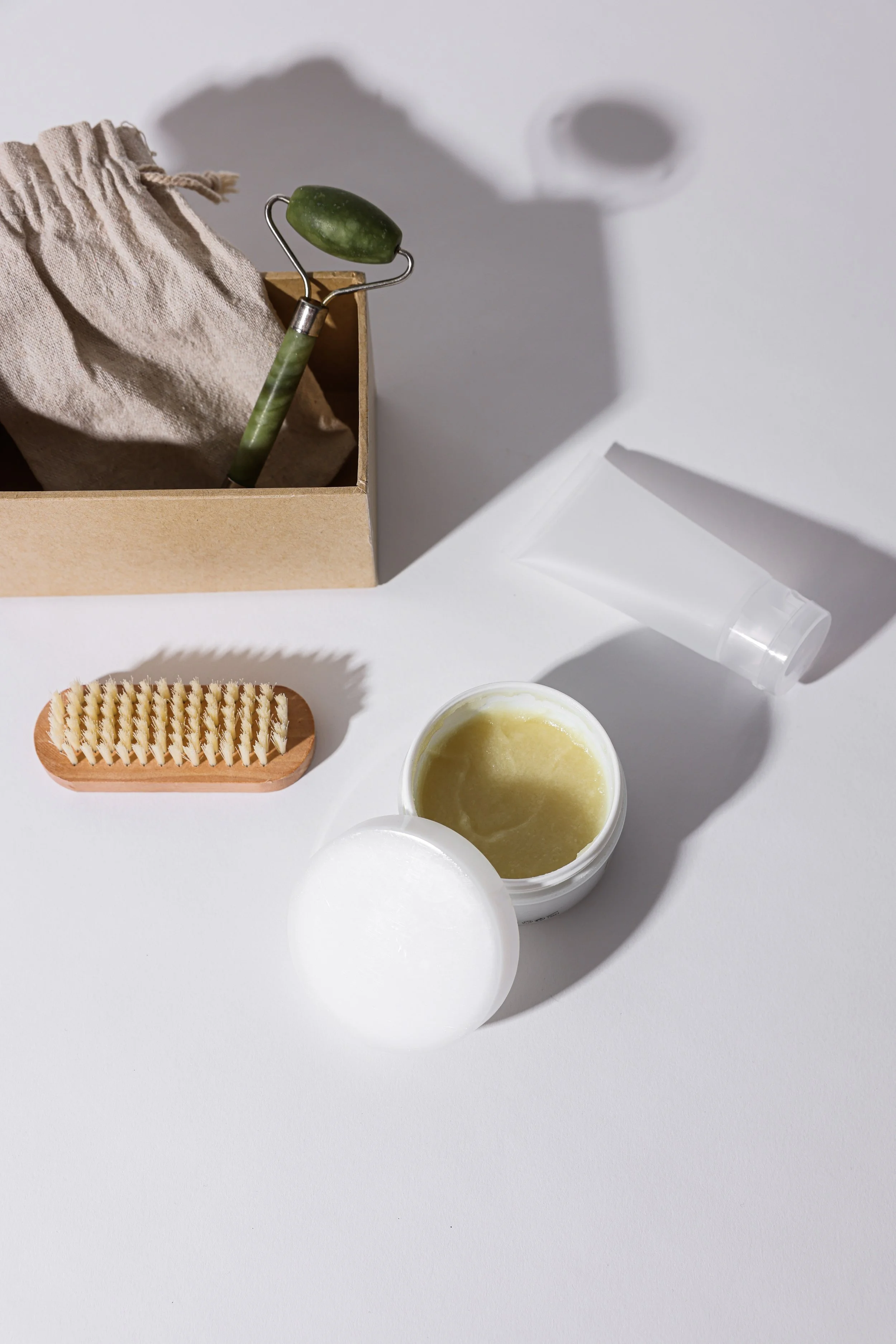 Skincare items on a white surface, including a small jar of balm with a white lid, a wooden nail brush, a clear plastic tube, and a cardboard box with a jade roller and cloth bag.
