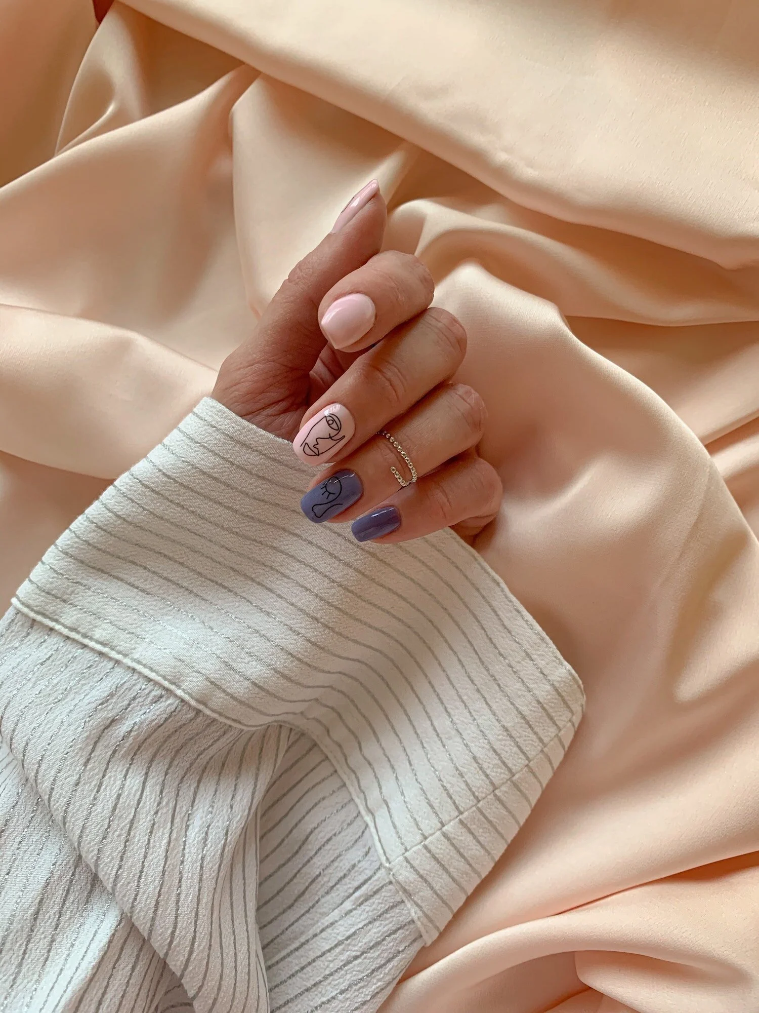 Close-up of a hand with painted nails holding a soft, peach-colored satin fabric. The nails are decorated with minimalist line art designs, two are pink with line drawings and one is purple with a line drawing. The person is wearing a striped fabric sleeve and a thin ring.