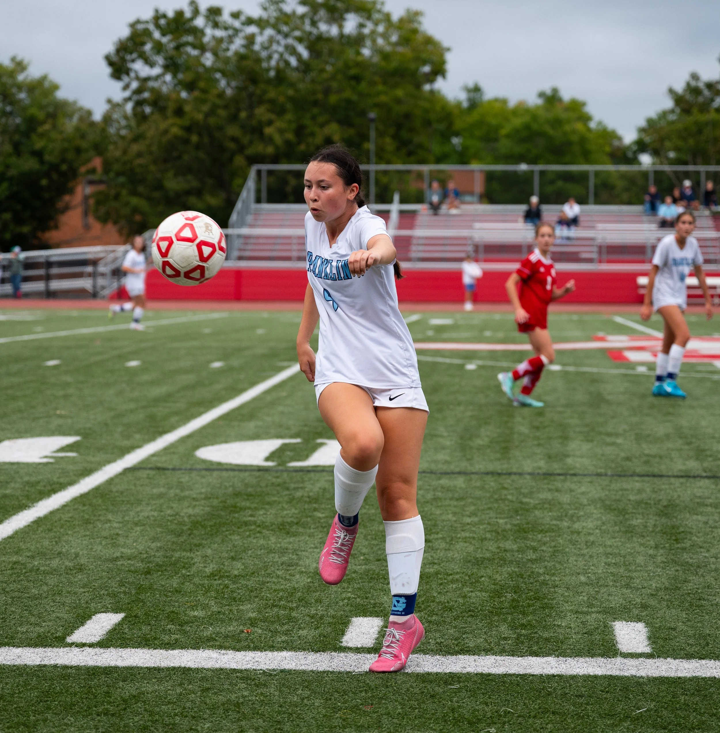 A young female soccer player in a white uniform is kicking a soccer ball on the field during a game. Other players and spectators are visible in the background.