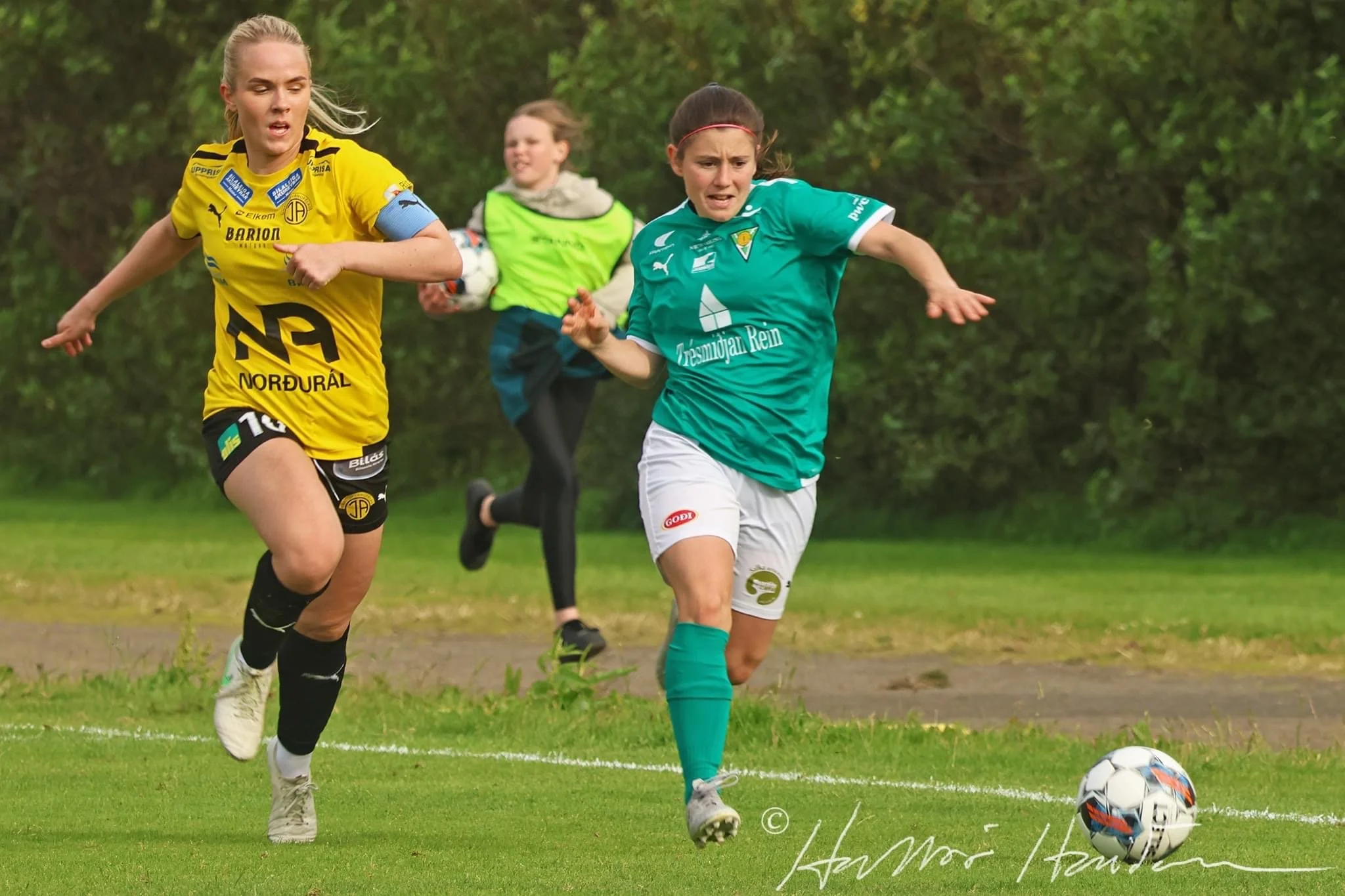 A young female soccer player in a white uniform is kicking a soccer ball on the field during a game. Other players and spectators are visible in the background.