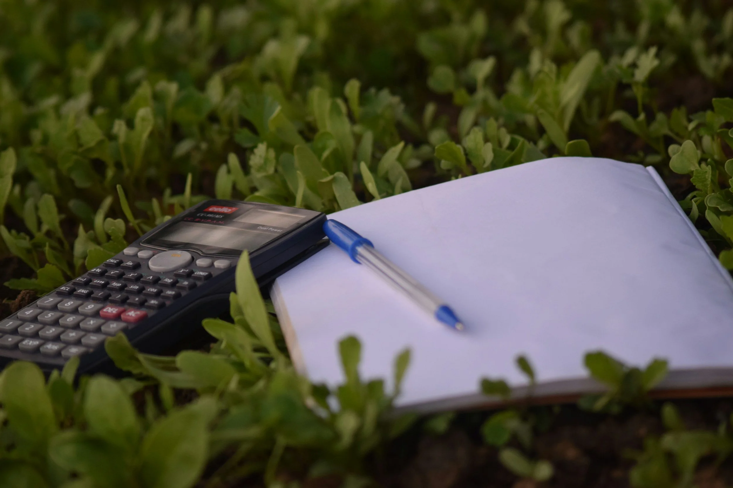 Calculator, notebook, and pen on green grass.