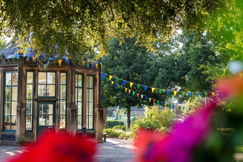 A stone building with big windows (the roundhouse in Burley in Wharfedale). It is sunny and there is colourful bunting, with trees in the background. In the foreground are colourful flowers