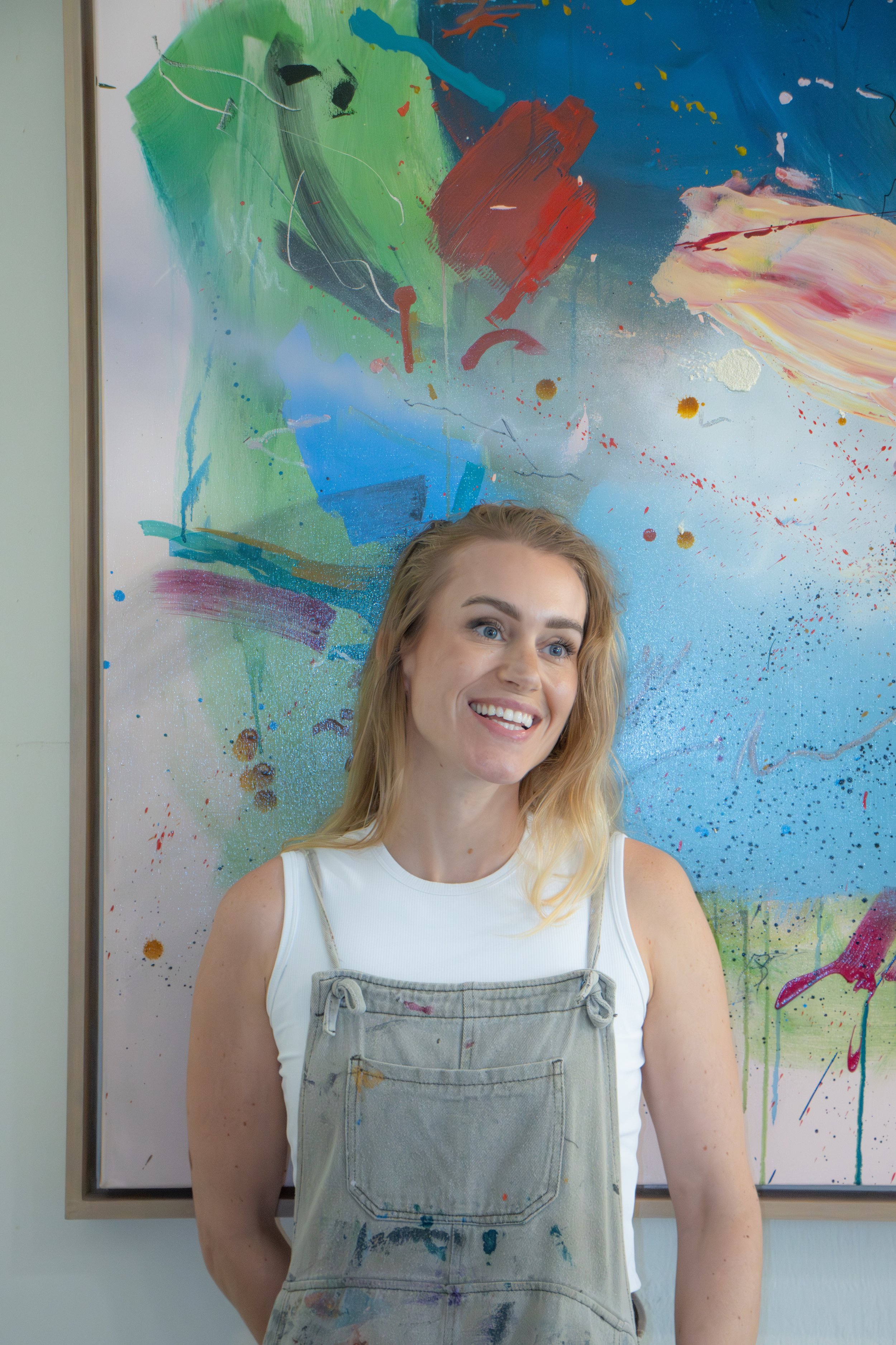 A smiling woman with blonde hair wearing a white tank top and a paint-stained apron, standing in front of an abstract colorful painting.