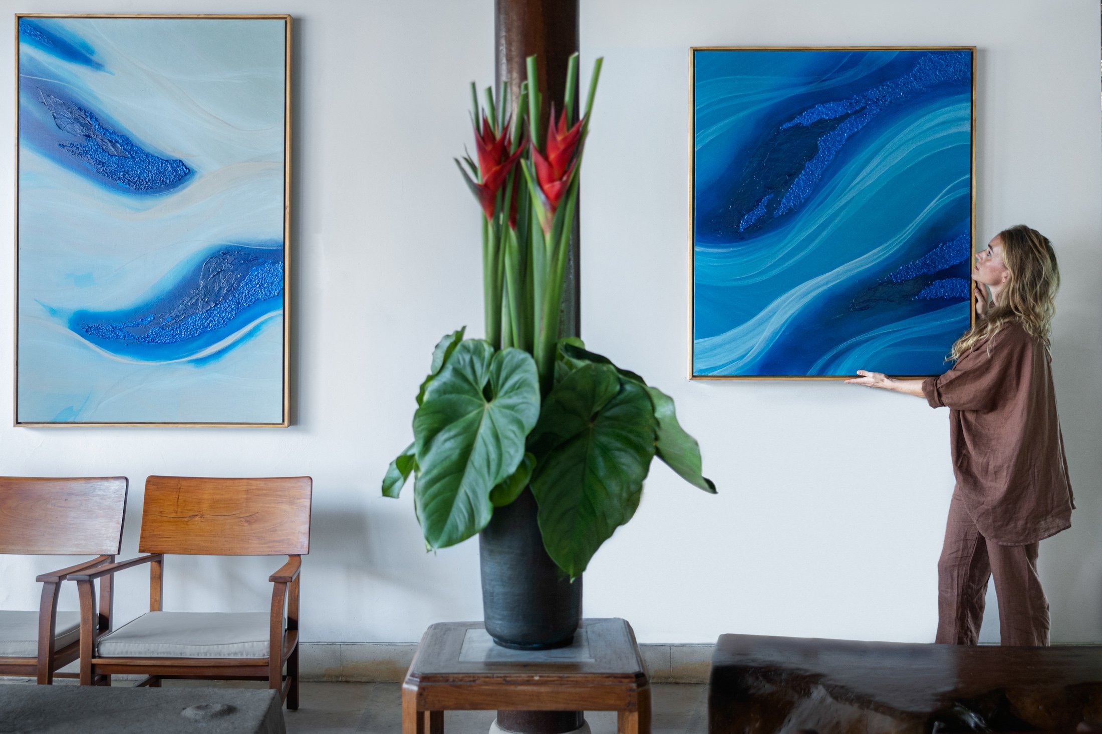 A woman with curly hair holding a large blue and black abstract painting in a gallery or art display space. There are two wooden chairs and a potted tropical plant with red flowers and large green leaves in the foreground.