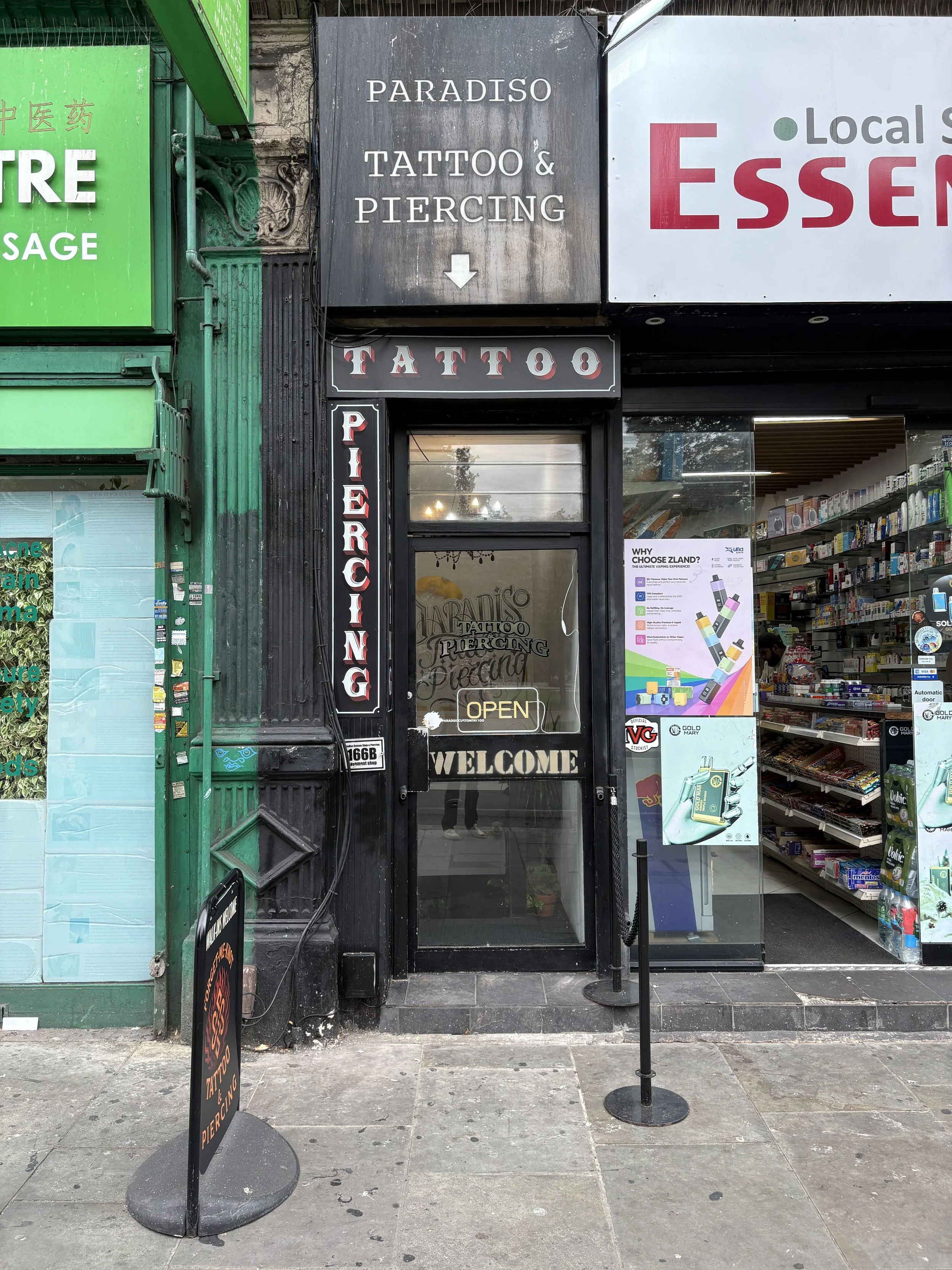 Storefront for Forget Me Knot Tattoo & Piercing, with signs on door and outside advertising tattoo and piercing services, and an open sign. Next to it is a pharmacy store with shelves visible through the glass.