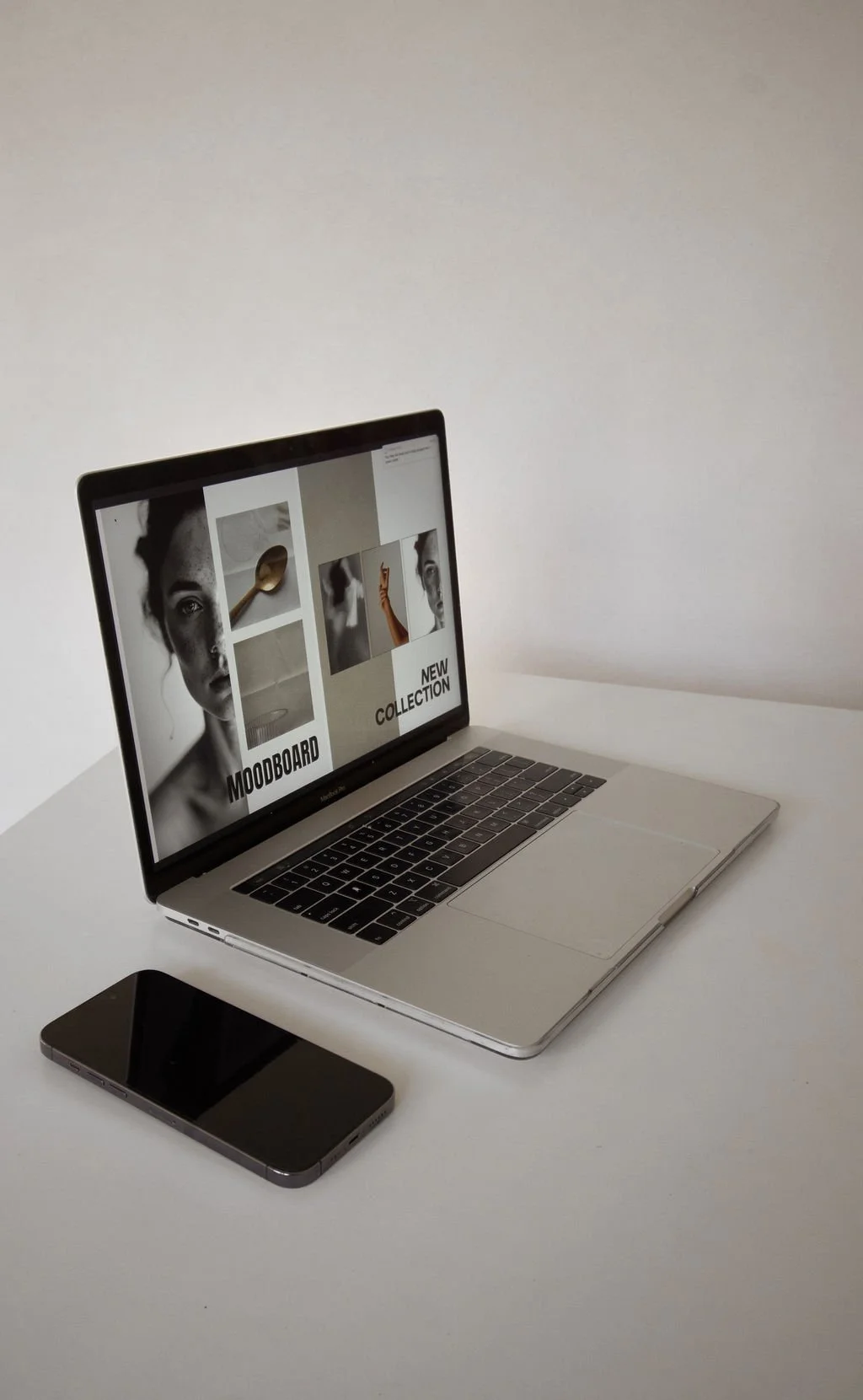 Laptop on a white table displaying a collage of black-and-white photographs and text that reads 'MOODBOARD' and 'NEW COLLECTION'; smartphone lies next to the laptop.