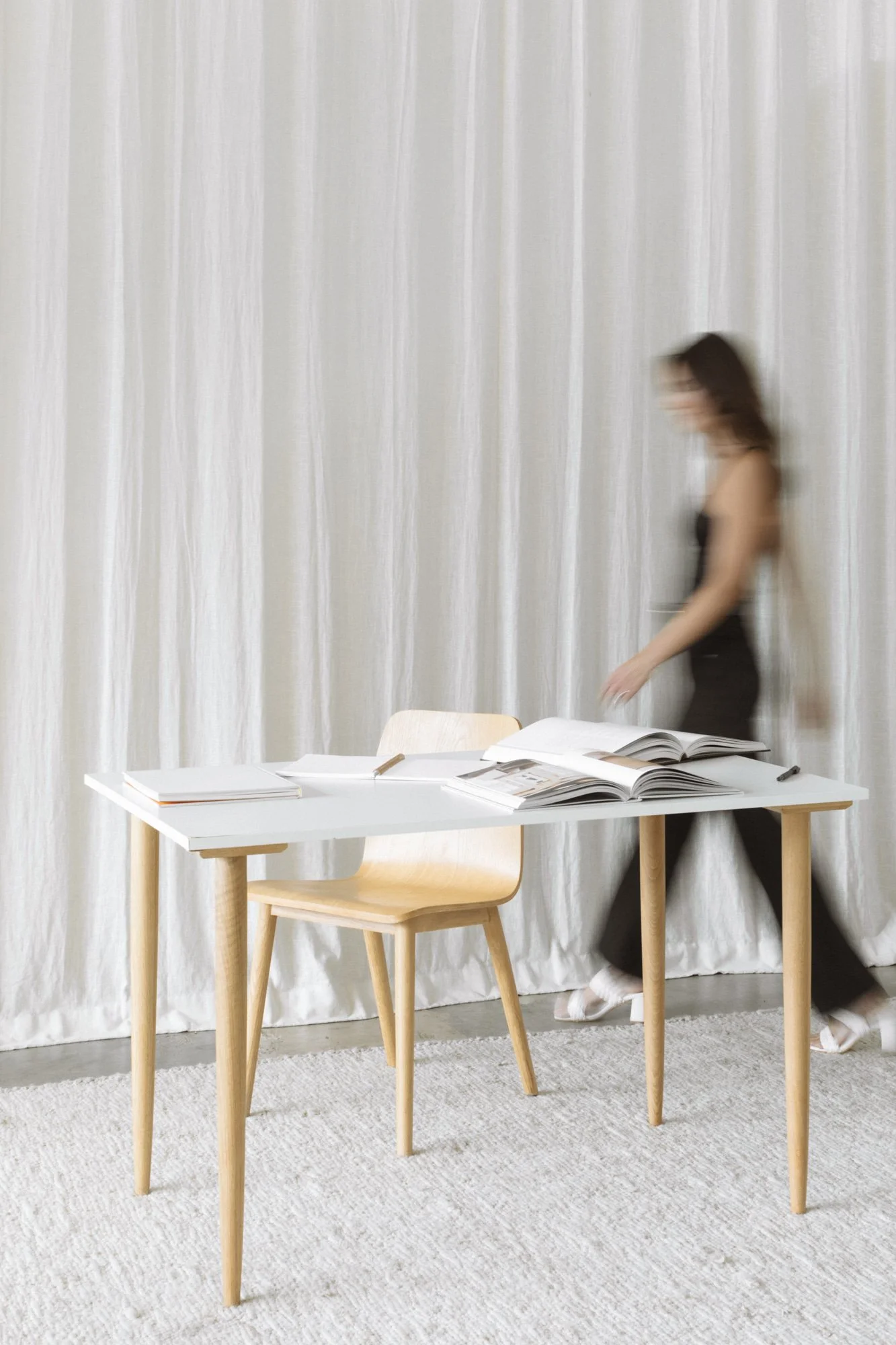 A woman in black walking past a white table with open books and writing materials, in front of white curtains.