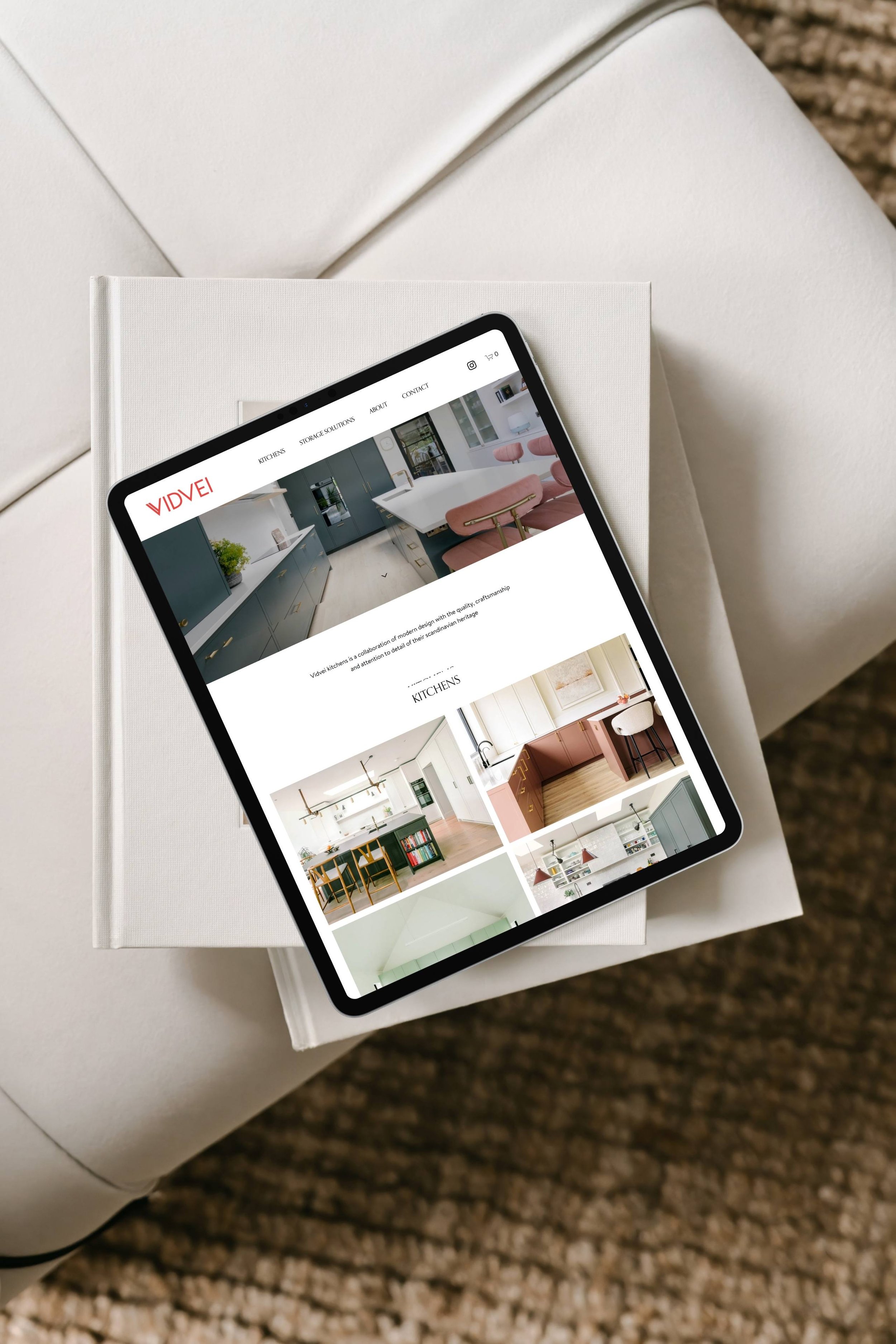 A tablet displaying a website with kitchen designs, resting on top of two stacked white books on a white upholstered chair with a woven carpet below.