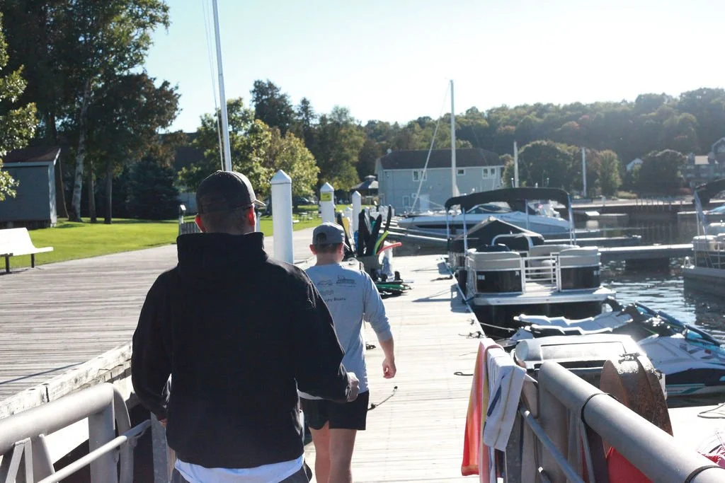 People walking on a wooden dock by a marina with boats, trees, and houses in the background on a sunny day.