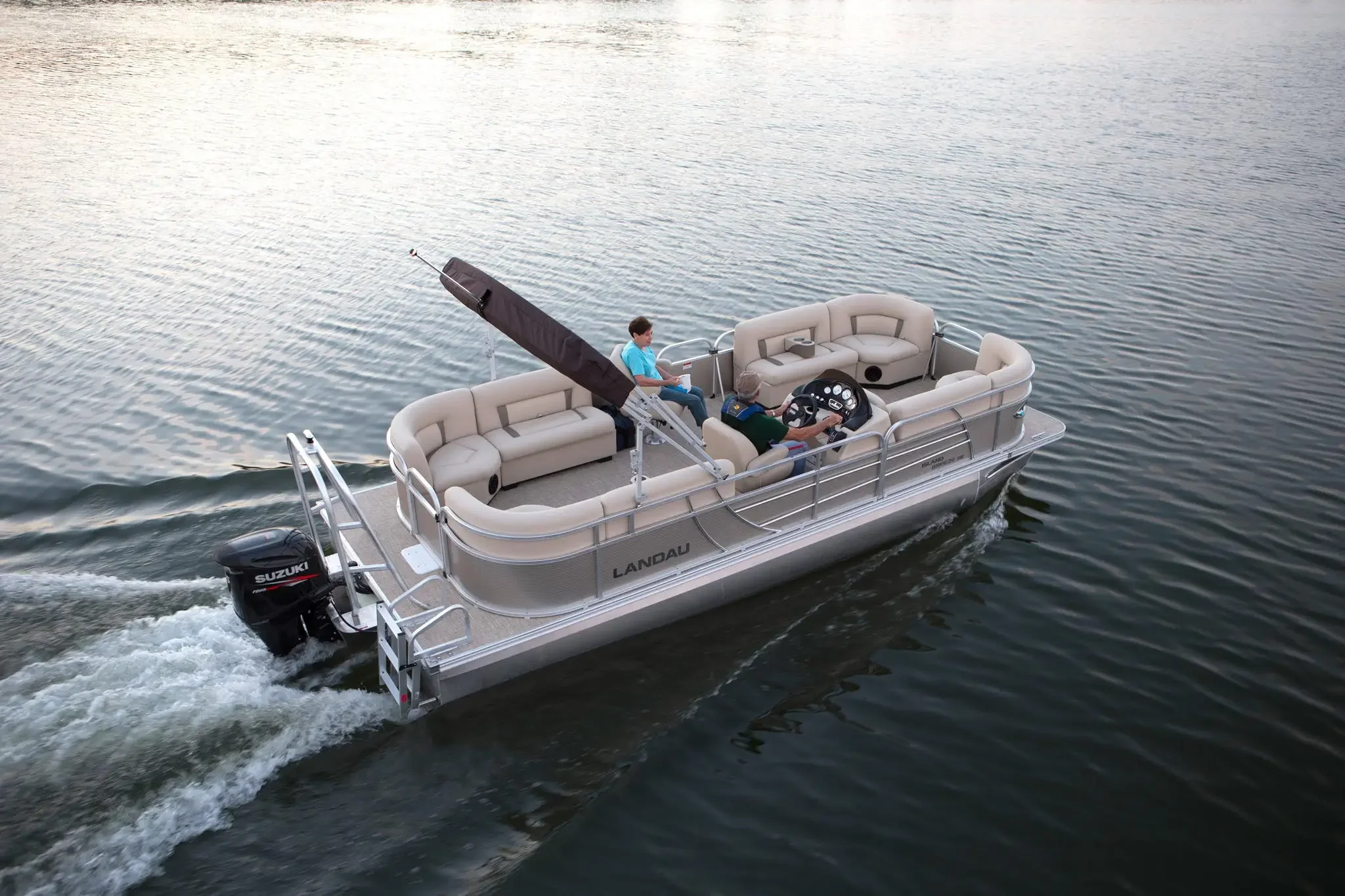 A pontoon boat with beige seating and a Suzuki outboard motor, cruising on a body of water. The boat has safety railings and two people are onboard, one at the steering wheel and another sitting near the center, enjoying the ride.