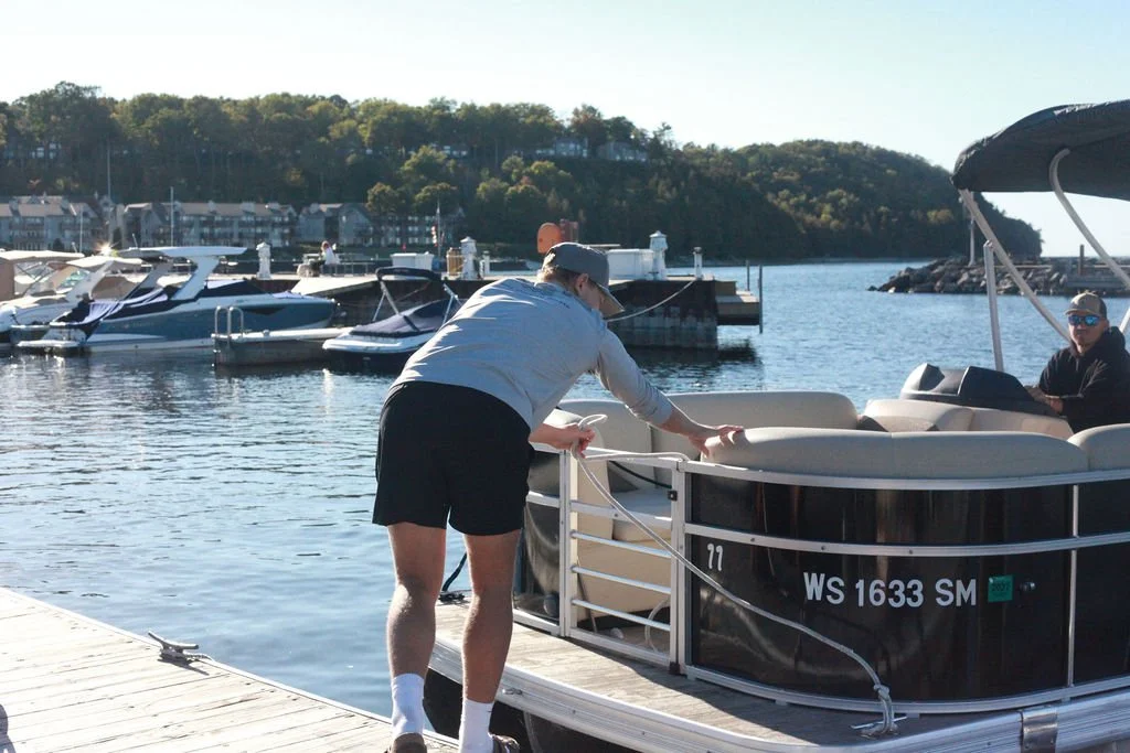 A person in shorts, a sweatshirt, and a cap is preparing a pontoon boat at a marina with multiple boats docked, water, and a tree-covered hillside in the background.