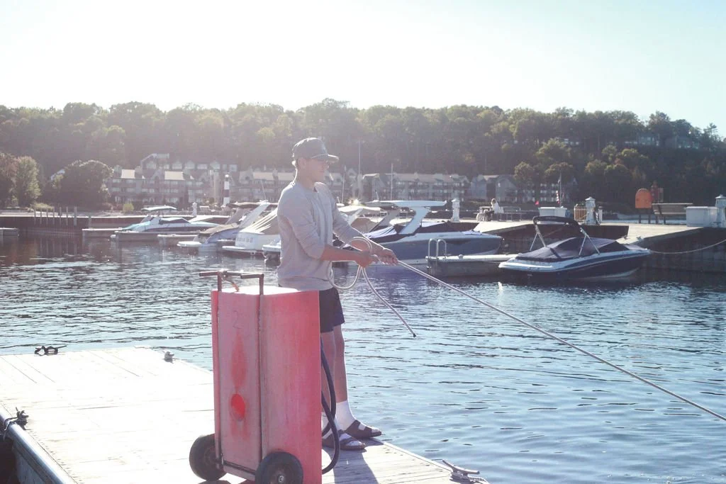 A person cleaning a dock with a power washer near boats in a marina, with residential buildings and trees in the background on a sunny day.