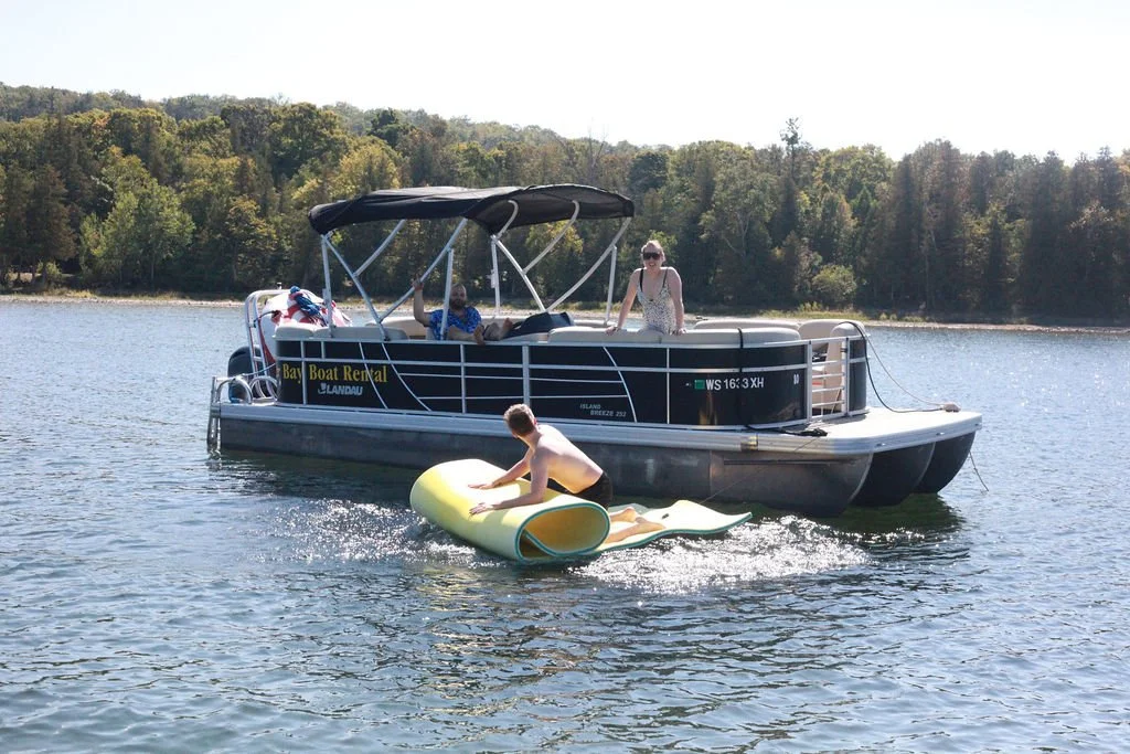 A boat on a lake with two people on board and a person on a floating mat in the water.