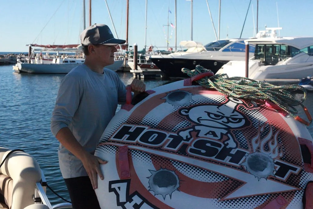 A person wearing a gray shirt and cap holding a large inflatable tube with a graphic design that says 'Hot Shot' and features flames and tire marks, near a marina with boats docked in the water.