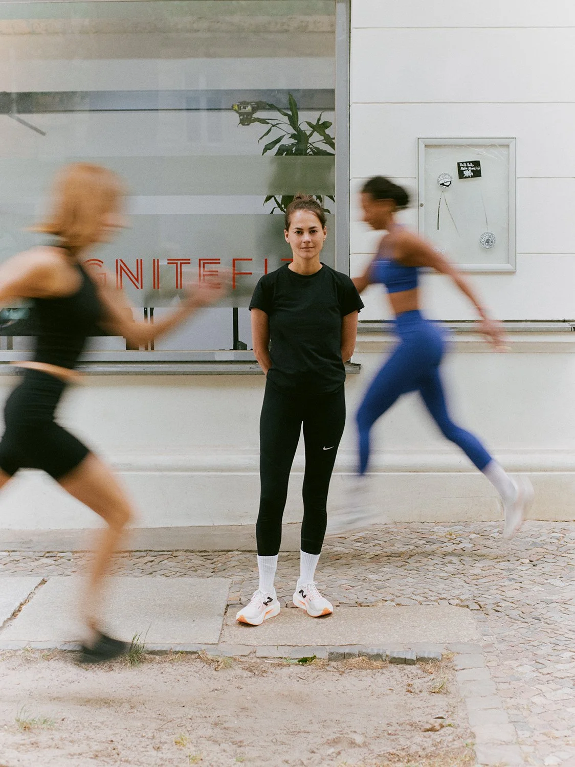 A woman stands still as two runners pass by in motion blur outside a gym, supporting wellbeing and mindful movement